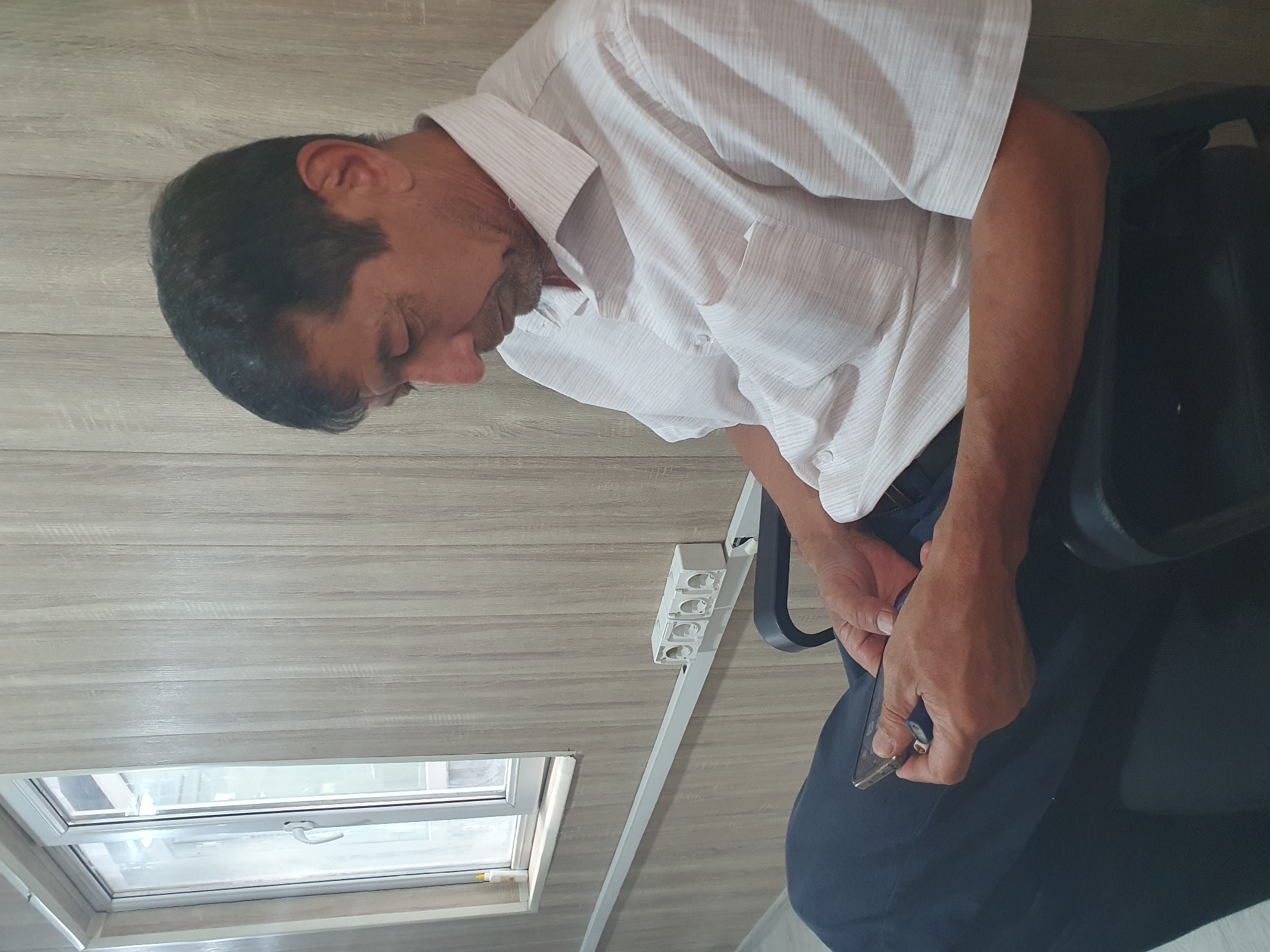 An adult male, appearing to be of Central Asian descent, sits in a black chair, looking down at a smartphone he is holding in his hands. He is wearing a light-colored, short-sleeved collared shirt and dark pants. The background consists of a wood-paneled wall with vertical planks and a window on the left side. A white electrical conduit runs horizontally along the wall, with a white outlet strip containing four receptacles attached to it. The scene appears to be indoors, possibly in an office or waiting room, located in Tashkent, Uzbekistan.