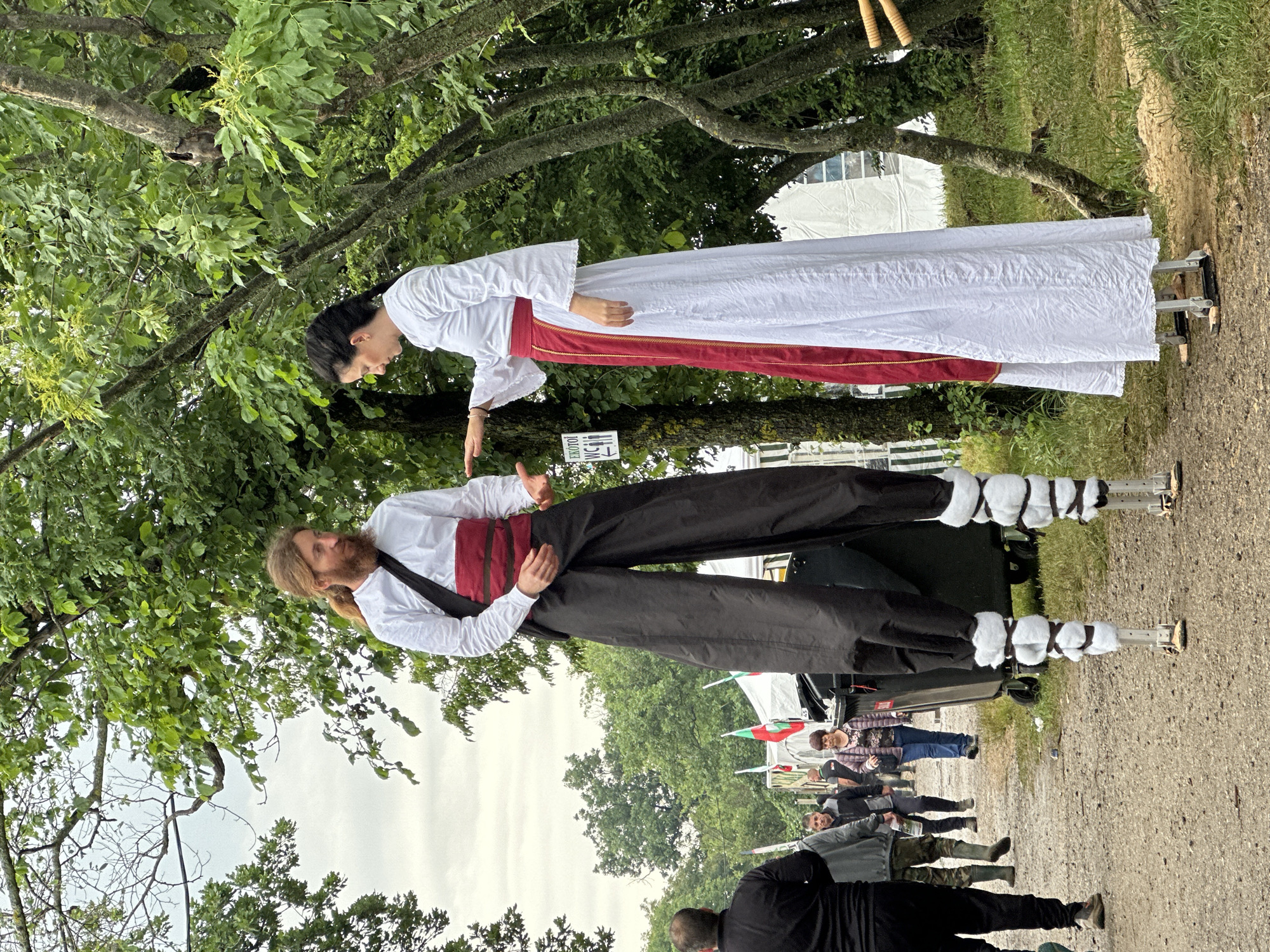 Two individuals on stilts in traditional Bulgarian costumes. A man is wearing a white shirt, black pants, a red sash, and white leg wrappings, supporting a dark haired woman in a white dress with a red sash. They are standing on a paved path, with trees overhead. A crowd of onlookers is visible in the background. The location appears to be outdoors. The context is the city of Lyaskovets, Bulgaria.