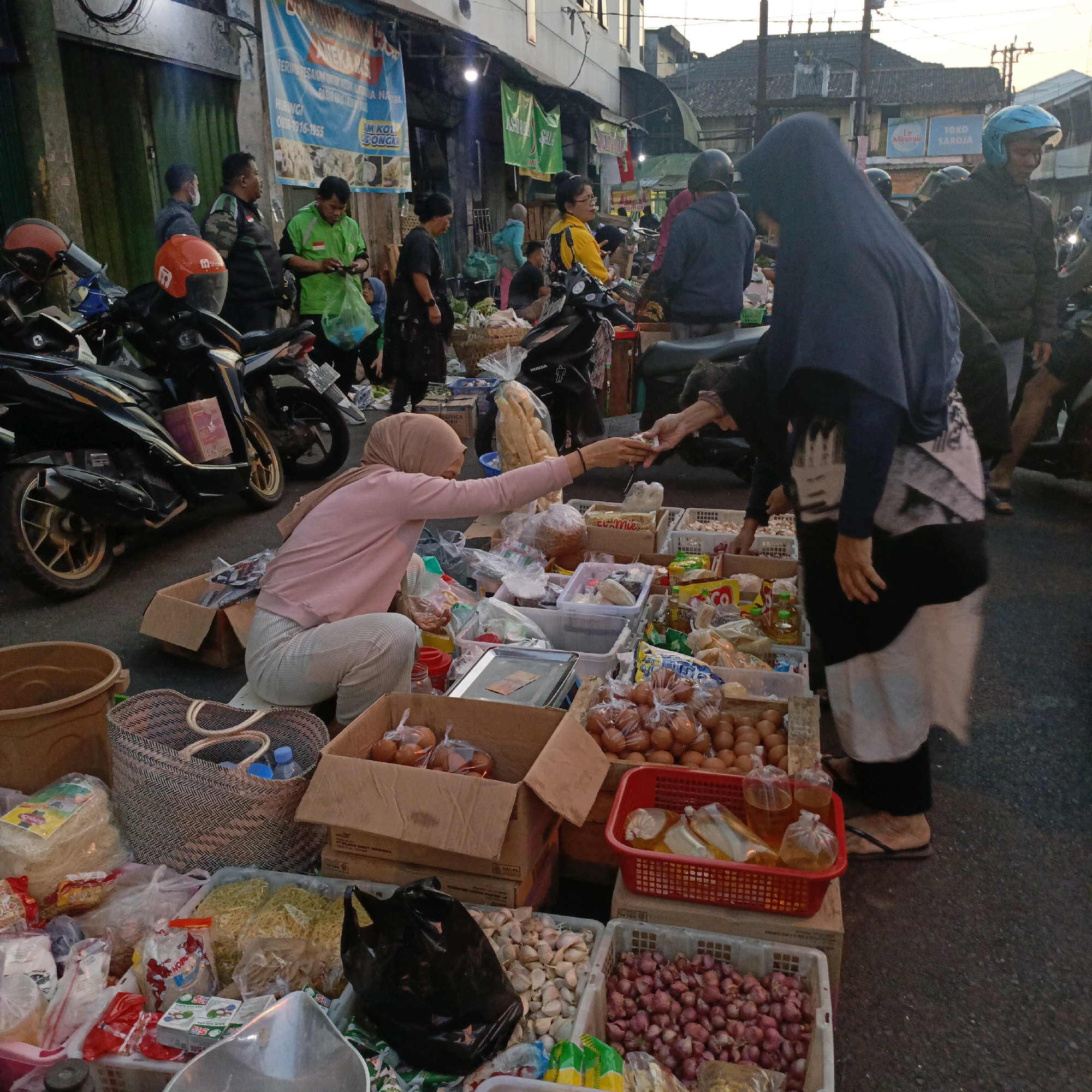 The image depicts a street market scene in Salatiga, Indonesia, likely on a late afternoon due to the soft, diffused lighting. The foreground is occupied by various food items and merchandise displayed for sale. A woman wearing a hijab and a light pink long-sleeved shirt and grey pants is crouched down, interacting with another person whose hands are visible reaching out to her. Boxes and baskets filled with eggs, shallots, garlic, noodles, and other packaged goods are arranged on the ground. Behind the stall, motorcycles are parked along the side of the street. In the background, the market extends, with more stalls, people, and buildings visible. Several individuals are seen wearing helmets, suggesting they may be vendors or customers arriving on motorcycles. A large blue banner with Indonesian text is prominently displayed on a building in the background. The overall atmosphere suggests a typical bustling marketplace environment.