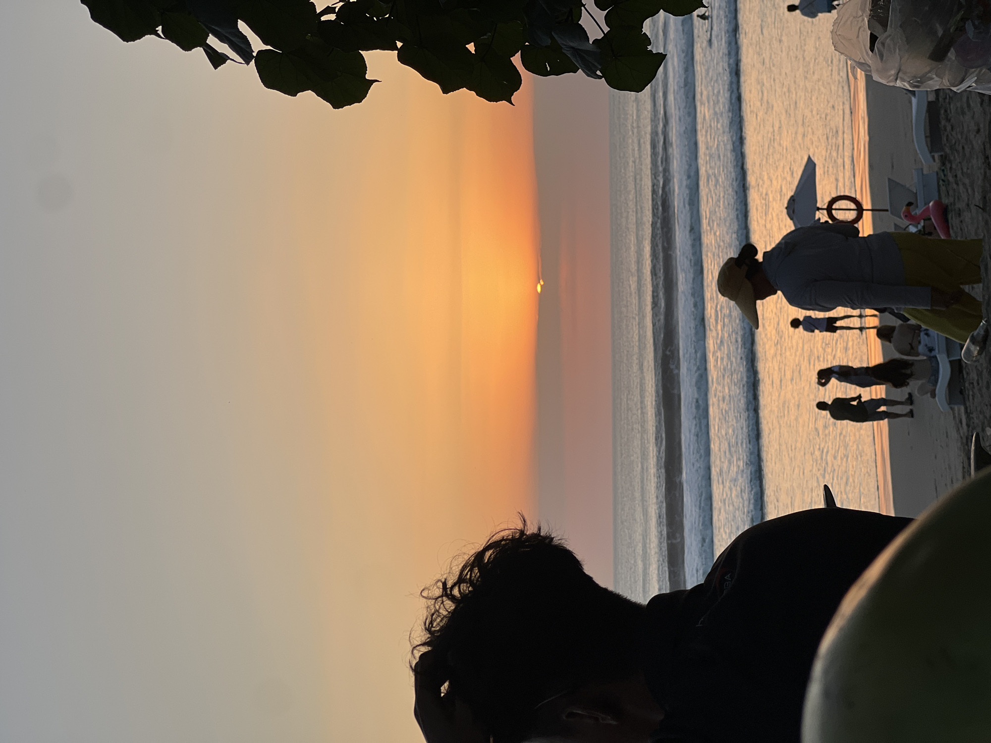 The image depicts a serene beach scene in Kuta, Indonesia, during sunset. The sky is a canvas of warm orange and yellow hues, with the sun partially visible on the horizon. The ocean is calm, with gentle waves lapping at the shore. In the foreground, a person with curly hair is seen in silhouette, touching their head. Further down the beach, several individuals are gathered, some walking along the shoreline, while others relax on beach chairs. Among them, a person wearing a wide-brimmed hat and yellow shorts is prominent. Beach umbrellas and a pink flamingo float add to the leisurely atmosphere. The scene is framed by lush green foliage on the right side of the image.