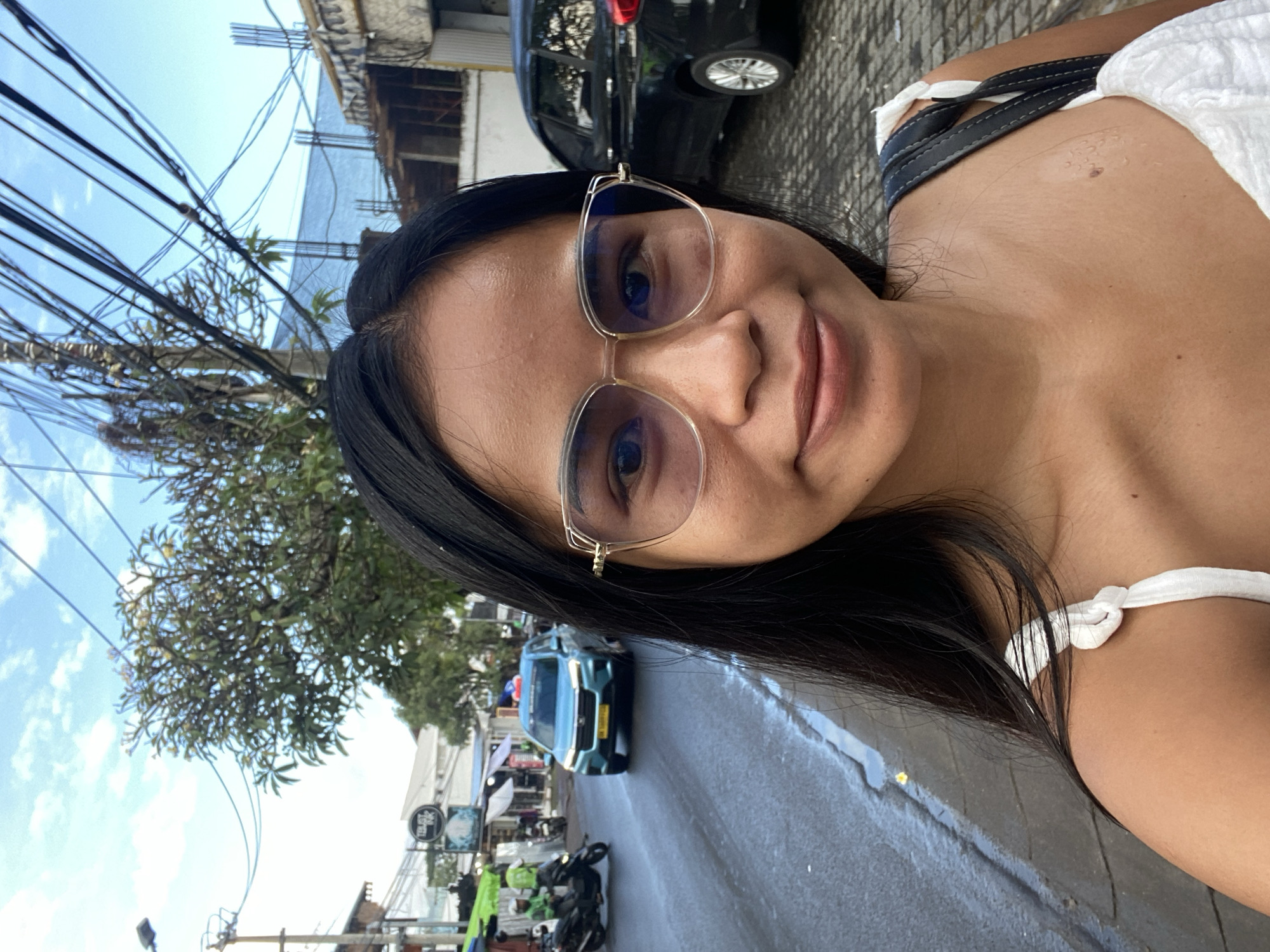 A daytime medium-shot selfie of a woman in a tropical urban environment in North Kuta, Indonesia. The woman has straight, dark hair, fair skin, and is wearing glasses with gold frames. She is wearing a light-colored top with straps. In the background, there is a street with a green car and a black car, as well as motorcycles parked along the side. A tree with lush green leaves is visible. Power lines crisscross overhead against a blue sky with scattered clouds. The street appears wet, indicating it may have recently rained. Other establishments and signs are also visible down the street.