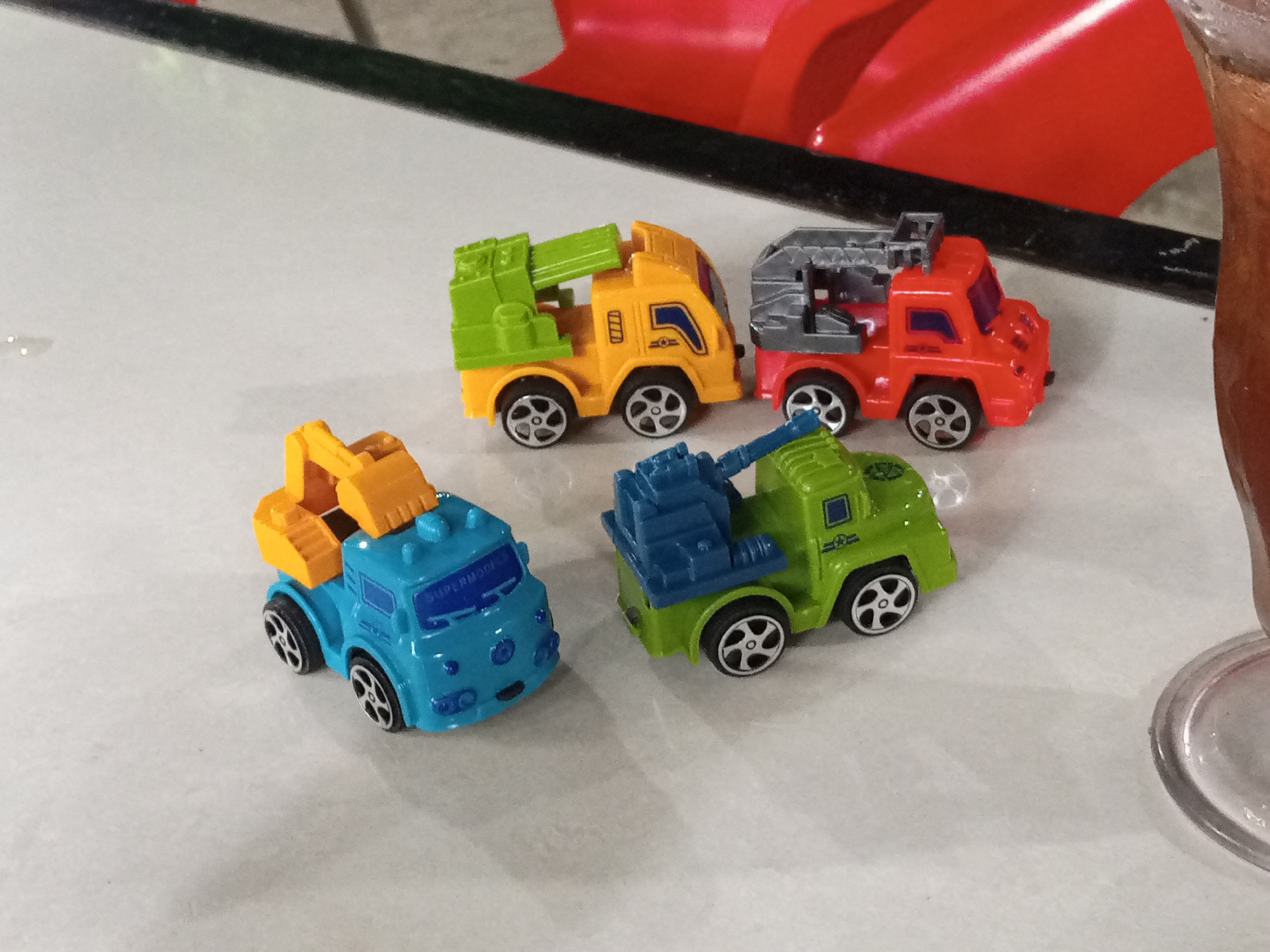 Four small toy trucks are arranged on a light-colored, speckled surface, possibly a table or countertop. The trucks are brightly colored: one is blue with a yellow excavator arm, another is yellow with a green crane, a third is red with a grey lifting mechanism, and the fourth is green with a blue crane. The blue truck has "SUPERMODELS" written on its windshield. The trucks appear to be lined up in a casual arrangement, with no specific activity or event depicted beyond their presence. The lighting suggests an indoor setting, and the reflective surface might indicate it's a glossy tabletop. A portion of a glass with a dark beverage is visible on the right side of the image, and a blurry red, curved object is in the background, possibly part of a larger item or decoration. The overall impression is a collection of toy vehicles displayed on a flat surface.