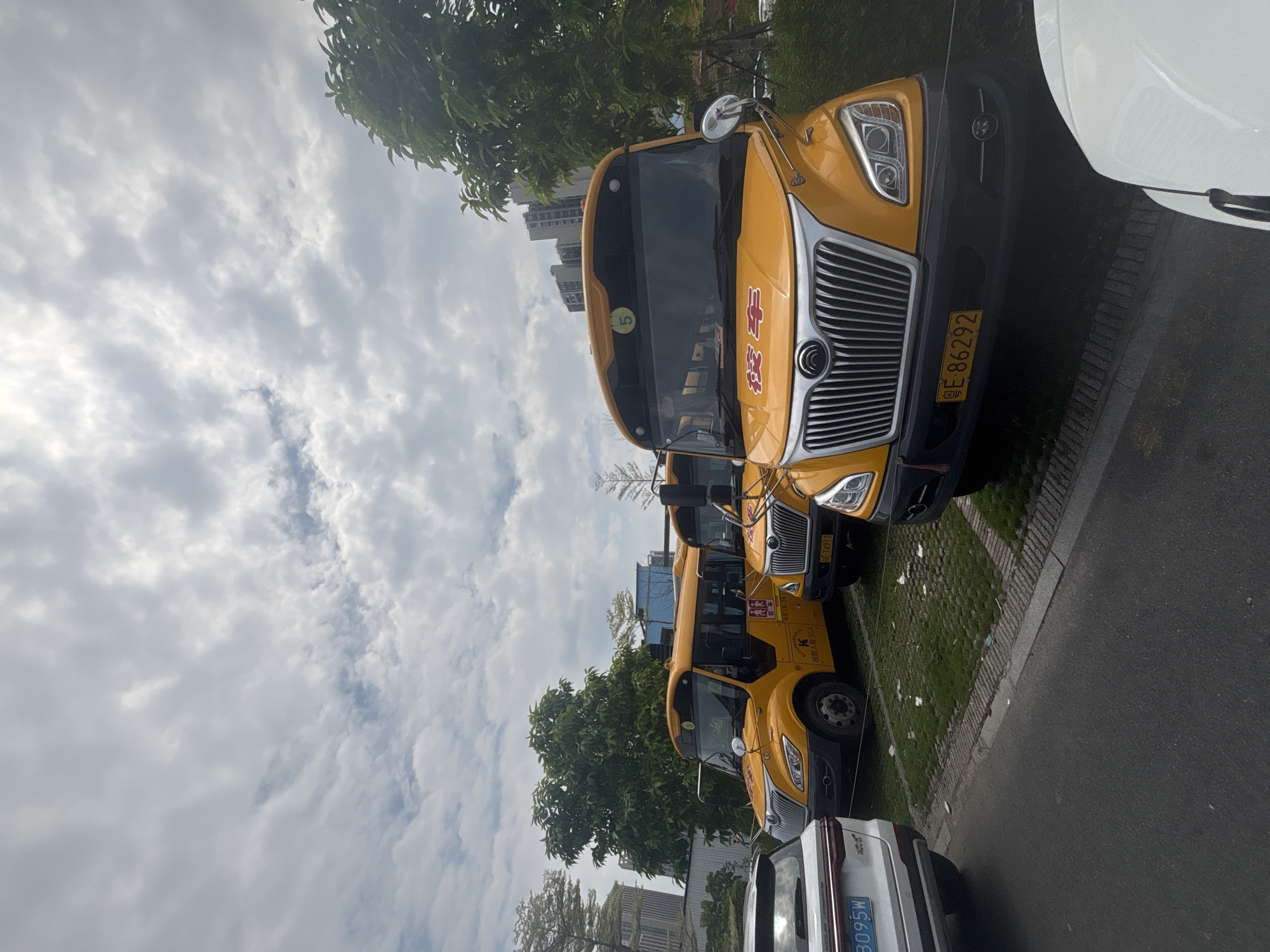 The image shows a row of yellow school buses parked in an outdoor lot, likely in the city of Nanhai District, China. The sky is overcast with visible clouds. In the foreground, a white SUV is parked to the left, and part of another white vehicle is visible on the far right. The buses are the primary subjects, with one prominent bus in the center displaying Chinese characters and a license plate that reads "E 86292". Another bus to the left has a license plate starting with "E". There are trees and buildings in the background, suggesting an urban or suburban environment. The ground is a mix of asphalt and paved parking spots with grass. No people are visible in the image.