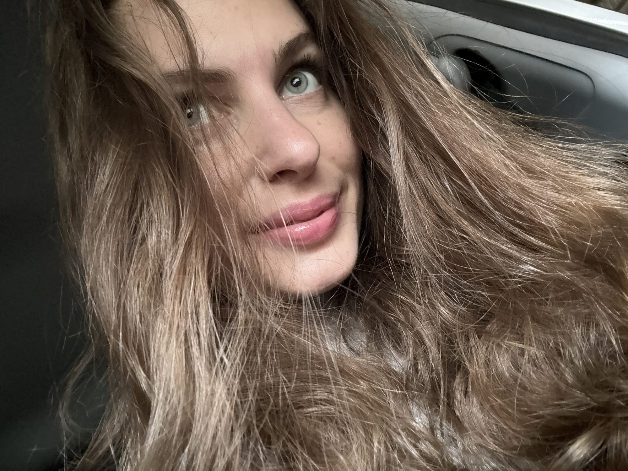 A fair-skinned woman with light brown hair and blue-green eyes is looking upwards in an interior setting. The woman has full, pink lips and appears to be sticking her tongue out slightly. The background is indistinct, but hints of a car interior are visible, including a handle and dark grey surfaces. The image is a close-up selfie. The city of Прикамский, Russia, cannot be verified as part of the context from the visual elements of the provided image.
