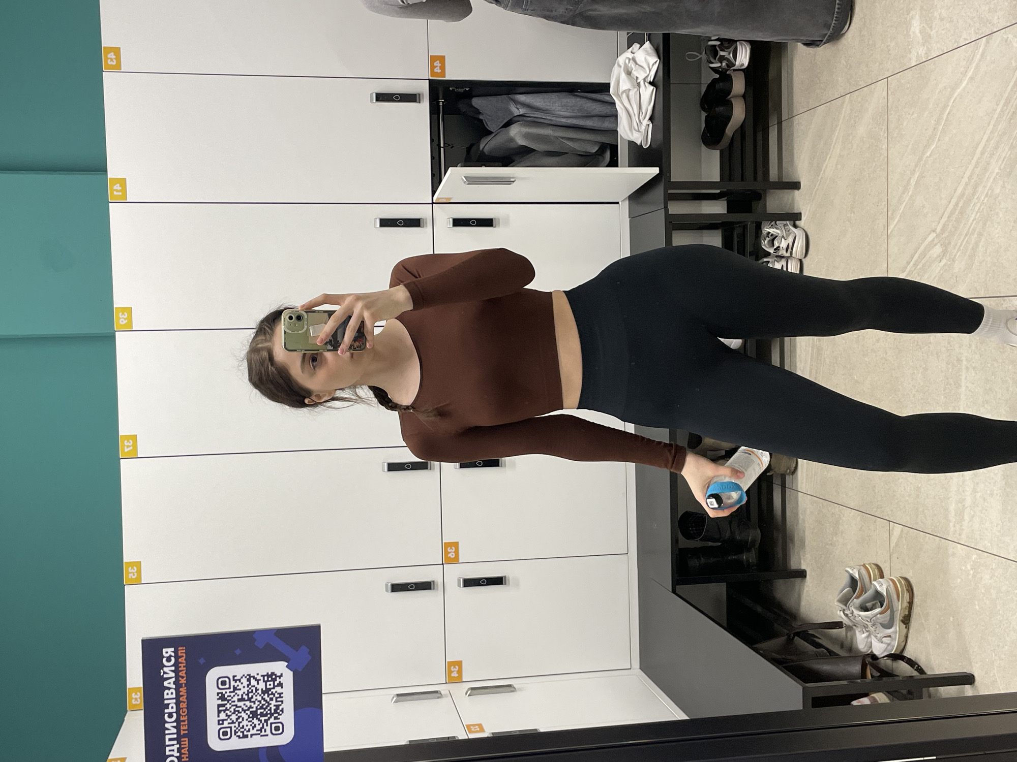 Here is a description of the image:
**Scene:** The image captures a young woman taking a selfie in what appears to be a locker room, potentially in a gym or sports facility.
**People:**
* **Subject:** A young woman dressed in activewear (a long-sleeved brown crop top and black leggings) is holding a smartphone to take a picture of herself in a mirror.
**Objects:**
* A smartphone is being held by the woman.
* A water bottle with a blue lid is held in the woman's other hand.
* Lockers lining the wall are white with yellow numbering, indicating locker numbers. There are lockers numbered 30, 32, 33, 34, and 39.
* A sign featuring a QR code and Russian text ("ОДПИСЫВАЙСЯ НАШ TELEGRAM-КАНАЛ!") is on the locker bank
* A shelf, likely for shoes or bags, holds various items: pairs of shoes, some clothing, and an open locker above it with a folded gray garment visible inside.
**Environment:**
* The flooring consists of light-colored tiles.
* The walls feature a combination of white locker fronts and a section of teal wall.
* The locker room has an overall clean and well-maintained appearance.
The Russian text "ОДПИСЫВАЙСЯ НАШ TELEGRAM-КАНАЛ!", which is on the QR code sign, translates to "Subscribe to our Telegram channel!". Considering the Russian text, it's probable that the location is in Russia, but without more details, it is difficult to pinpoint the specific city.