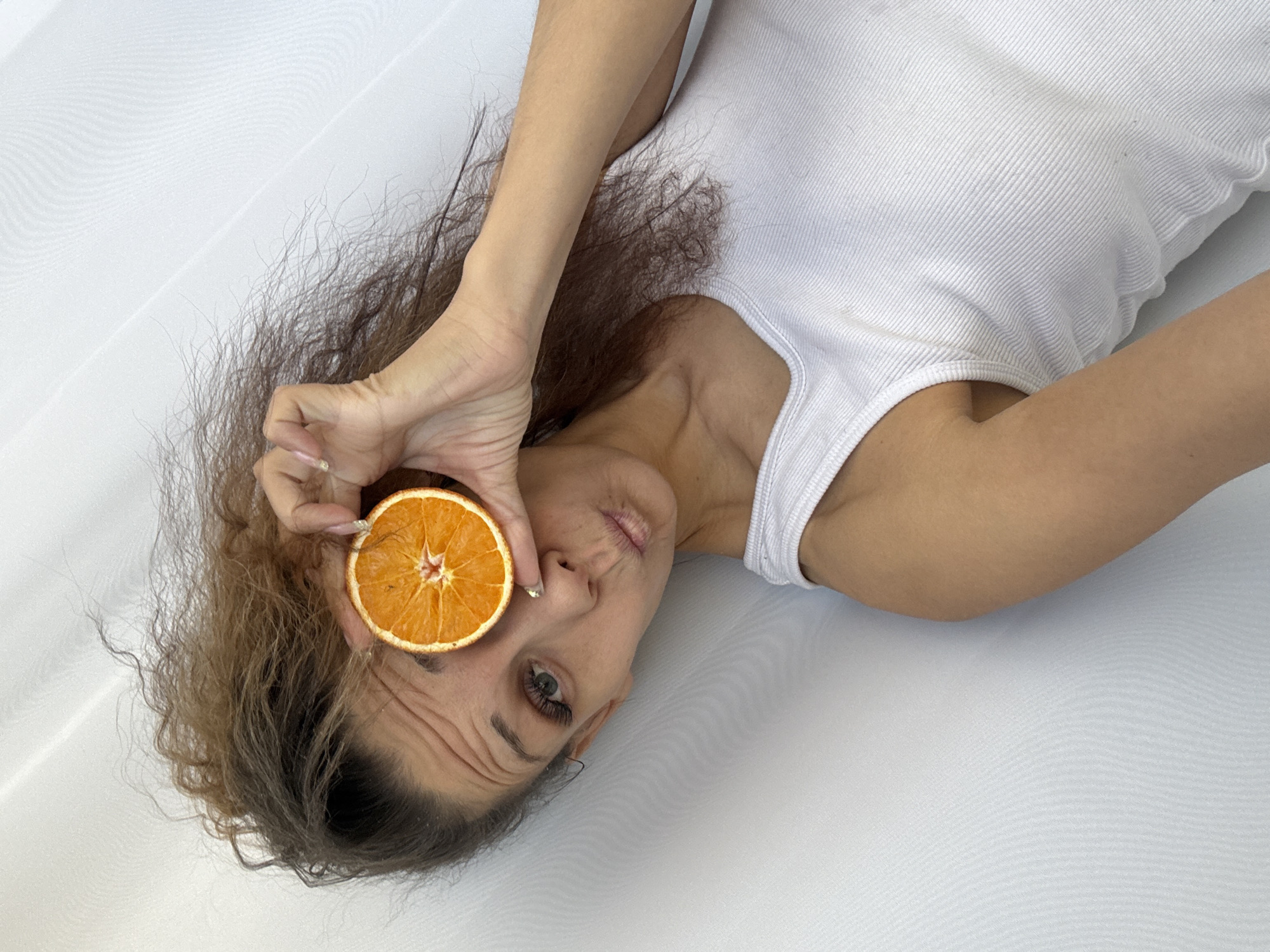A woman with brown curly hair is lying on a white surface, presumably a bed sheet. She is wearing a white tank top. She holds a dried orange slice over her left eye. Her face is tilted slightly towards the camera, and she is looking at the lens. The image appears to be taken from a high angle, looking down on the woman. There is no indication that this image was taken in Rusanovo, Russia.