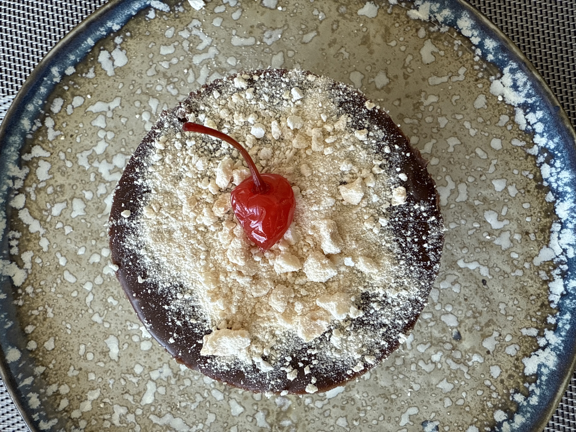 This overhead, close-up image features a single serving of dessert artfully placed on a decorative ceramic plate. The dessert is a dark, round cake or tart, generously covered with a light-colored, granular topping that appears to be a mix of fine powder and small, irregular crumbs. A single, glossy bright red maraschino cherry, complete with its stem, rests prominently in the center of the topping.
The plate itself is round and has a unique, rustic design. Its surface displays an mottled pattern of light beige and off-white speckles against a muted, earthy brown background, giving it an aged or artisanal appearance. The rim of the plate is bordered with an irregular band of dark blue and grey, adding to its handmade aesthetic.
A dark grey or black woven placemat is visible underneath the plate in the background, particularly on the left and upper right sides, suggesting an indoor dining setting. The lighting is even and soft, highlighting the textures of the dessert and the plate. There are no people visible, and no text is present in the image. The overall composition presents an appealing dessert ready for consumption.