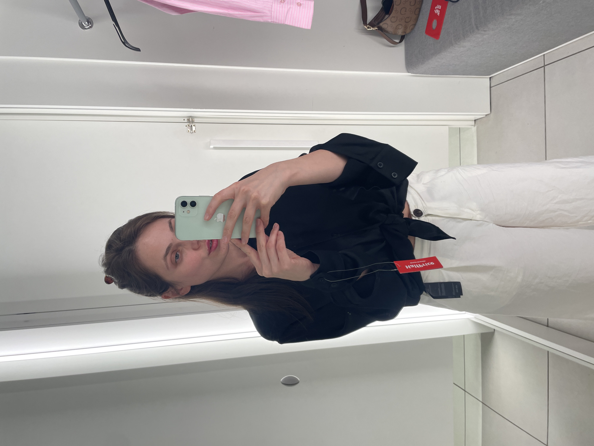 A woman is taking a mirror selfie in a fitting room. She is wearing a black tie-front blouse and white wide-leg pants. She is holding a mint green smartphone in her right hand. A red clothing tag is visible hanging from her blouse. In the background, a pink striped shirt is hanging on a rack, and a brown and tan monogrammed purse is resting on a gray bench. The fitting room has white walls and a gray tiled floor. The location is a fitting room in a clothing store.