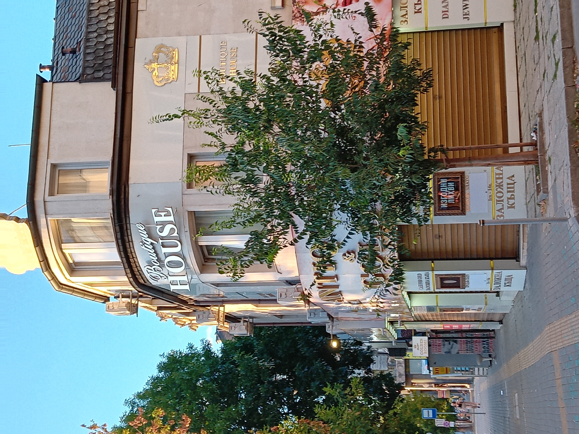 The image depicts a street scene in Varna, Bulgaria, under a clear blue sky. A multi-story building with a rounded corner dominates the foreground. The building's facade is adorned with signs, including "Boutique HOUSE" in large silver lettering and a golden crown emblem. Several air conditioning units are mounted on the exterior walls. A young tree with dense green foliage stands in front of the building.
To the left of the building, a sidewalk with a tactile paving strip runs alongside the street. A bus stop sign is visible on the sidewalk. A woman in casual clothing is walking away from the camera on the sidewalk. Various shops with shuttered storefronts line the street. Signs for "GOLDEN ROOMS," "CASH FOR GOLD," and "DIAMOND JEWELRY" are visible on some of these shops. The street is paved with interlocking concrete blocks. The overall impression is of a commercial street in an urban setting.