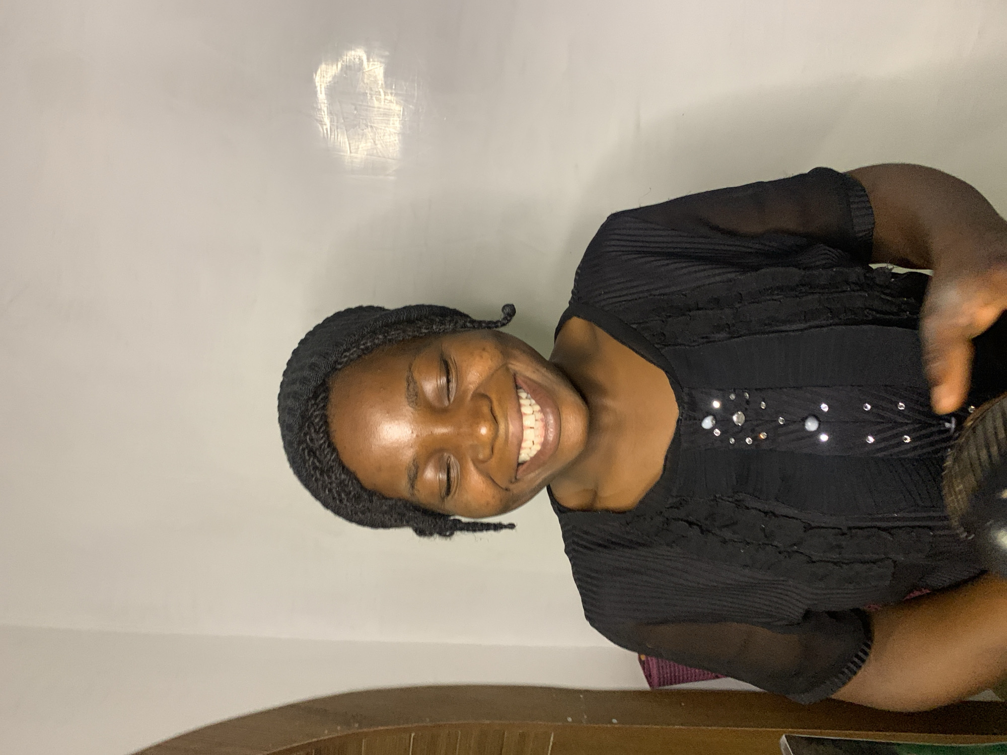 A young Black woman with dark skin and braided hair, wearing a black dress with decorative embellishments, smiles with her eyes closed. She is positioned in the foreground against a plain, light-colored wall. The environment suggests an indoor setting, possibly in Jos, Nigeria, given the context of the request. A portion of a wooden door frame is visible on the left side of the image. Her posture and expression convey happiness or amusement.
