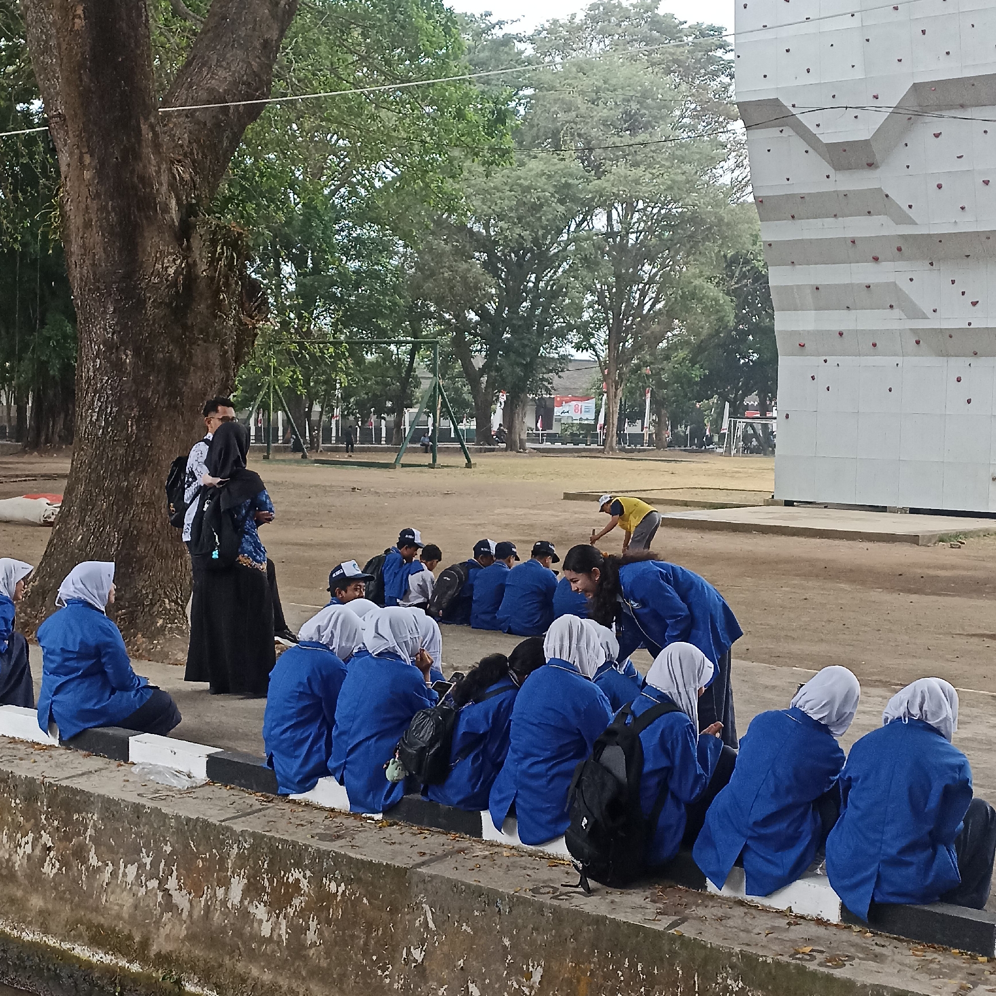 In Magelang, Indonesia, a group of students in blue uniforms and headscarves are seated on a concrete ledge near a large tree. A climbing wall stands to the right of the frame, and a playground structure is visible in the background. The students appear to be taking a break from an activity, with some looking at their phones and others conversing. A man and a woman, also in uniform, stand near the tree. The ground is dirt, with patches of grass and fallen leaves. The sky is overcast.