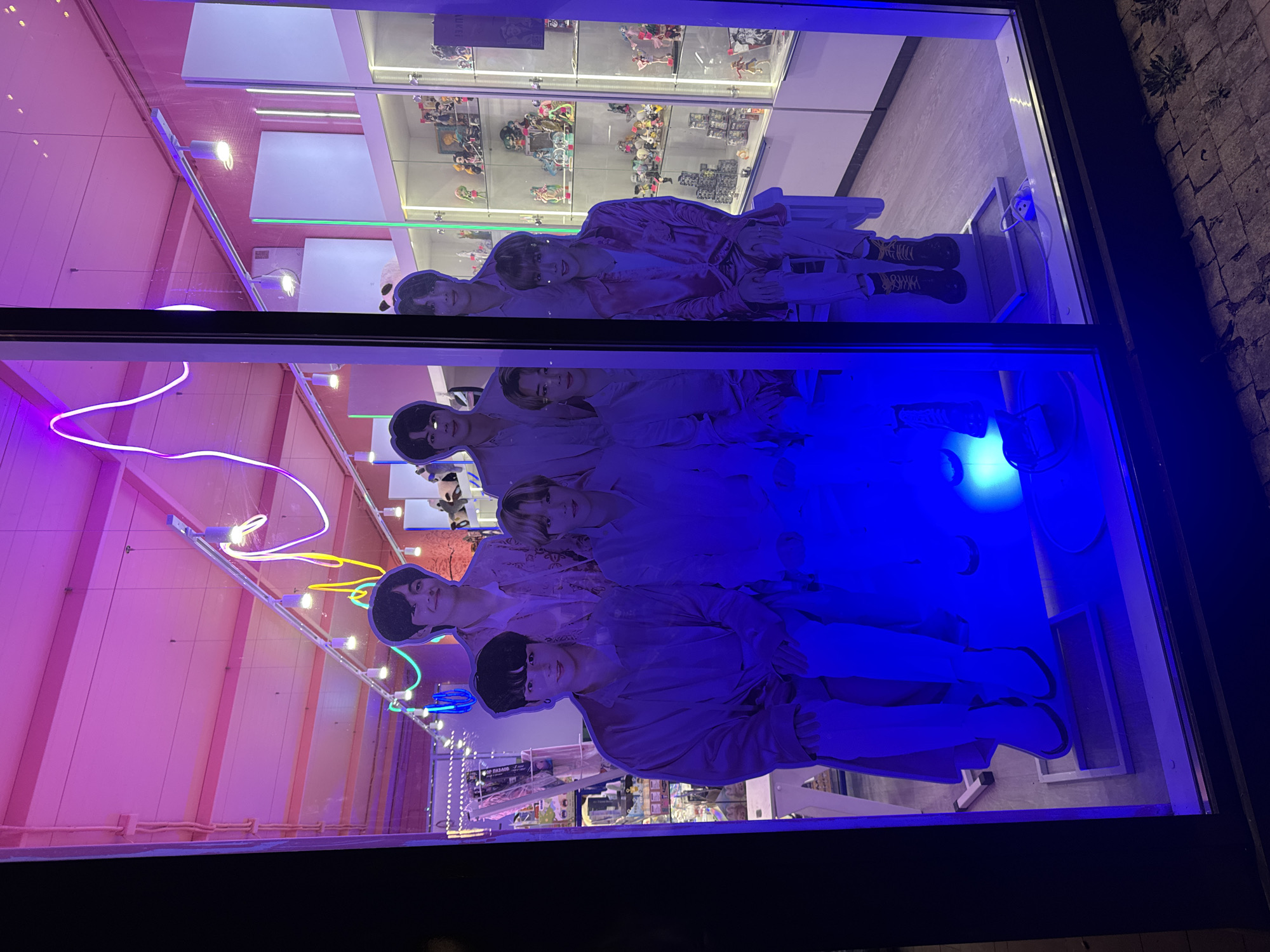 The image captures a shop window display. Inside, several life-size cardboard cutouts of the K-pop group BTS are arranged. Display cases filled with figurines and other merchandise are visible in the background. The window reflects the surroundings, including neon lights and the interior of what appears to be a retail space. The location is identified as the city of Unknown, Unknown.