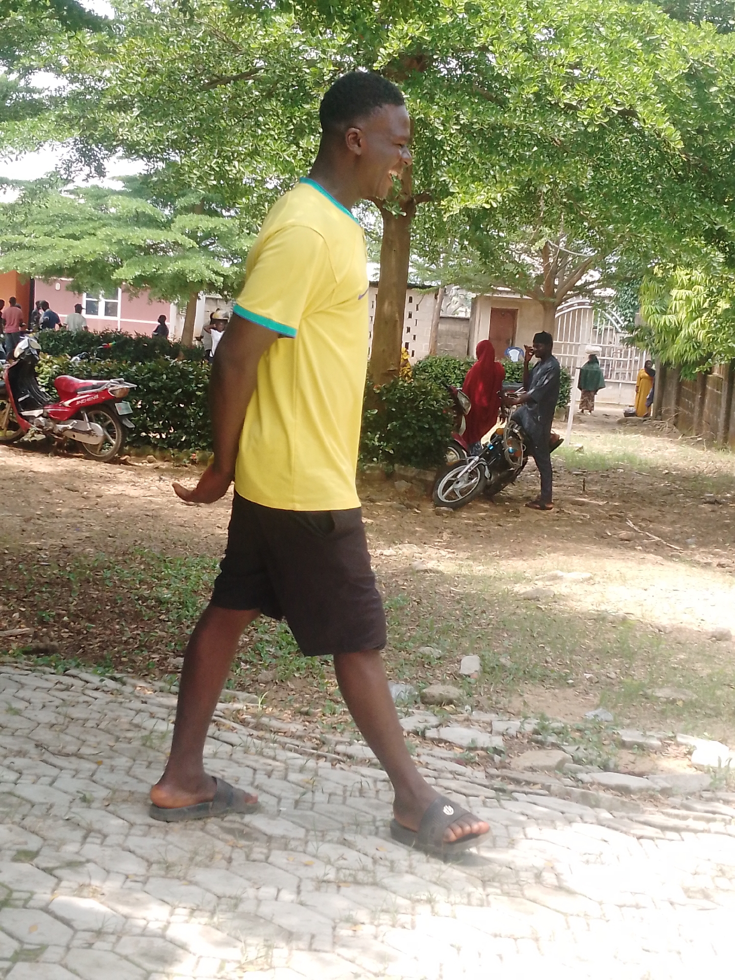 A side profile of a Black male wearing a yellow t-shirt and black shorts is walking on a paved walkway. He is looking to his right and smiling. Behind him, a red motorcycle is parked next to a hedge. Further in the background, several people are gathered near a building. The environment appears to be outdoors with trees and foliage. The scene is likely in Akwanga, Nigeria.