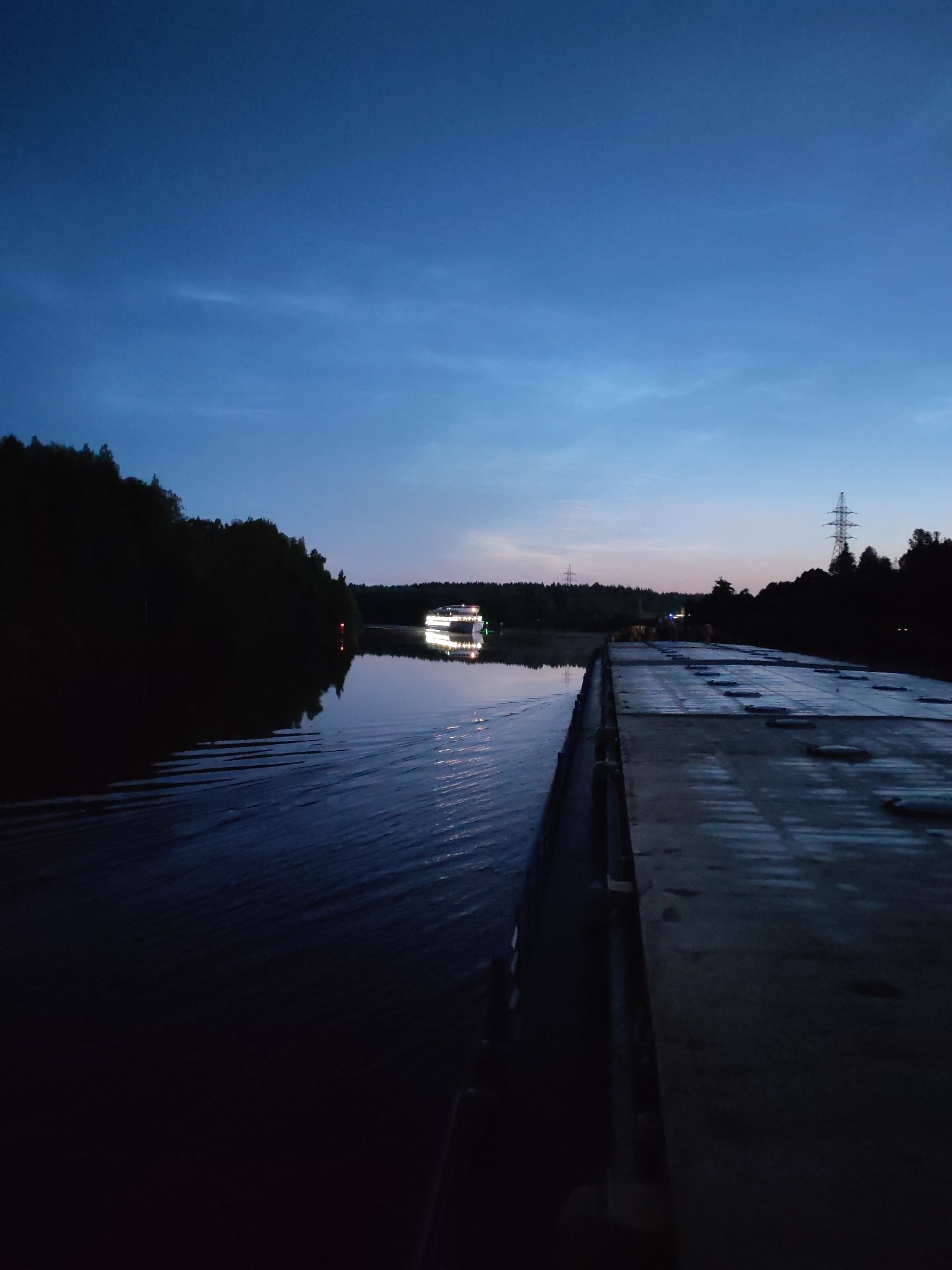 The image shows a body of water, likely a river or canal, at dusk. On the left side of the frame, a dense line of dark trees forms the bank. In the distance, a white boat with illuminated windows is traveling on the water, its reflection visible on the surface. On the right side of the frame, a long, flat, wooden structure, possibly a dock or barge, extends into the scene. The sky is a deep blue, transitioning to lighter shades near the horizon, with a few wispy clouds. In the far distance, on the right side of the horizon, a tall, latticed electrical pylon is visible. The overall impression is a tranquil, twilight scene in a natural or semi-natural environment. The location is stated to be Depo, Russia.