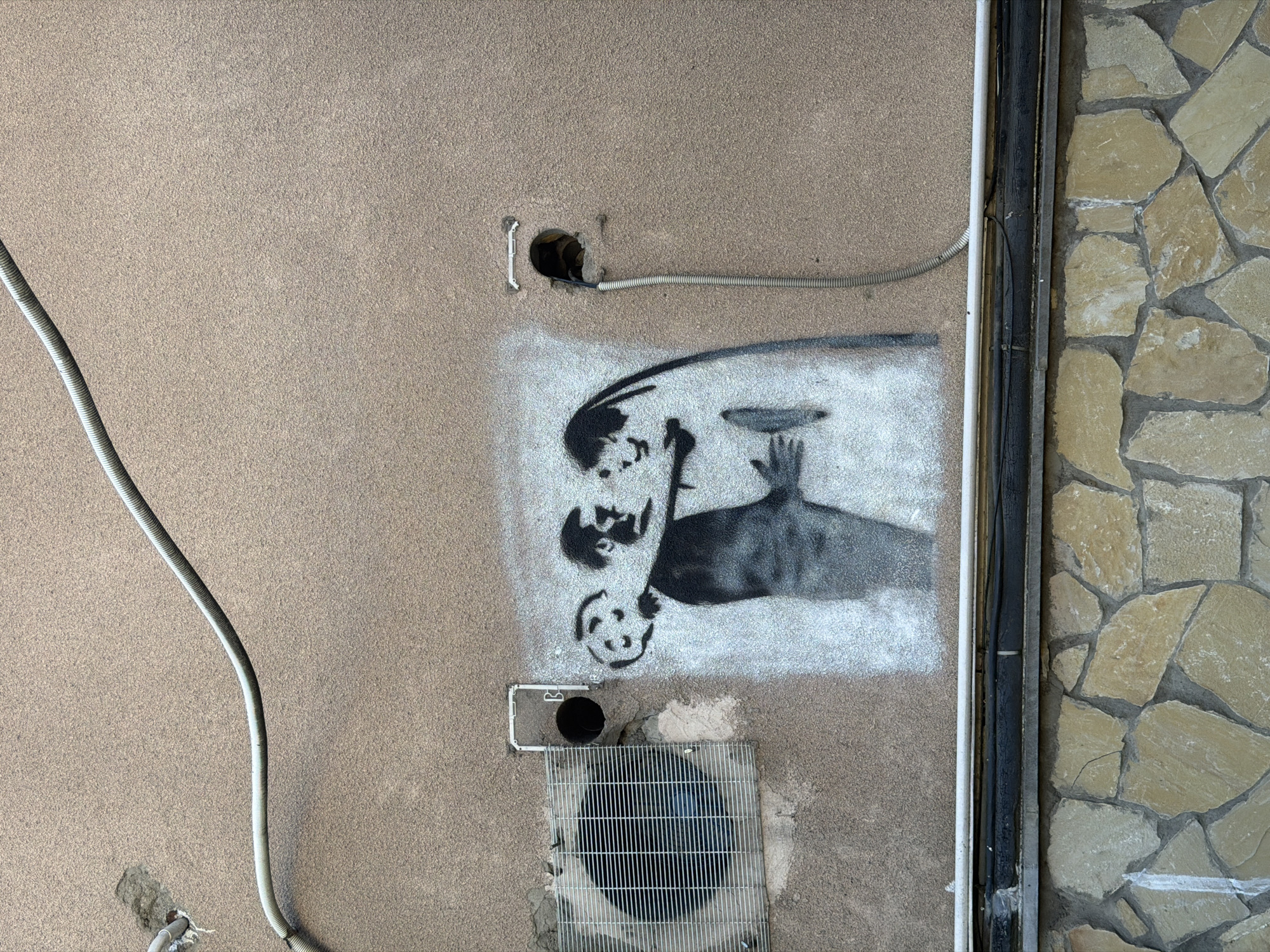 A section of an exterior wall in Tbilisi, Georgia, displays a tan stucco surface with a spray-painted Banksy-esque image in black and white. The image depicts three figures: one lying face down with a hand extended toward a plate or disc balanced precariously above; a second figure bending over the first as if to assist; and a third figure at the bottom with a smiley face. The artwork seems to reference the act of balancing something, potentially alluding to challenges and fragility.
Above the artwork are two conduit lines running horizontally across the wall. One protrudes from an open hole; the other runs parallel to the top of the artwork. Below the artwork, a metal grate covers a ventilation hole in the wall. To the right of the stucco wall, a column of irregularly shaped light-colored stones creates a border, separated from the stucco by a vertical gray structure. The scene is devoid of people. The overall impression is of an urban environment with a mix of artistic expression and functional utility.