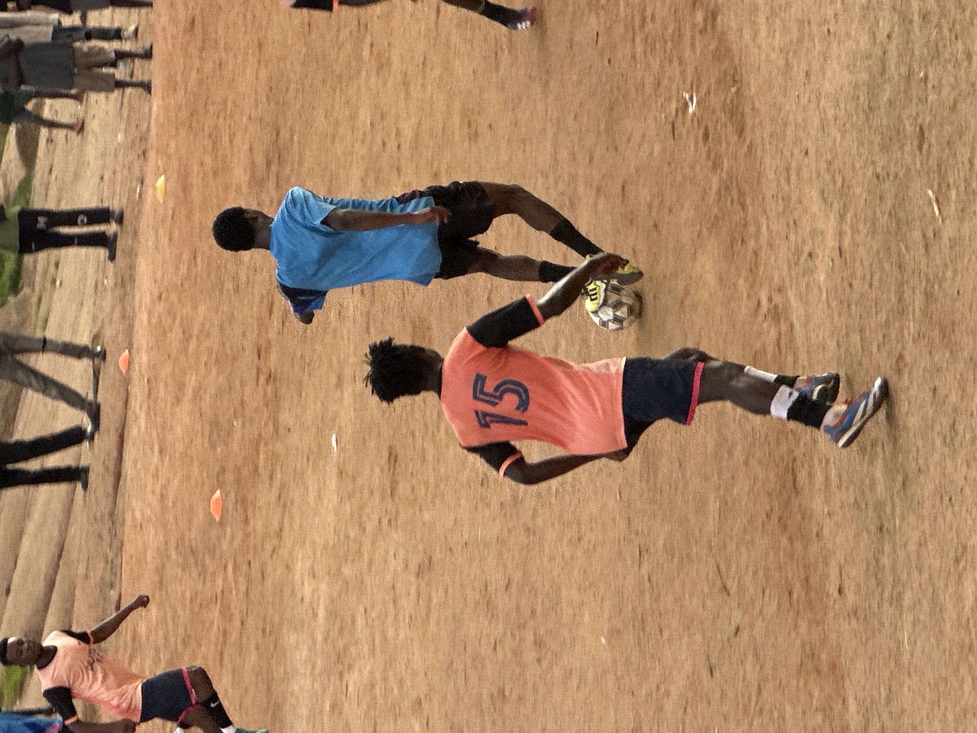 This image depicts a group of young men playing soccer on a dirt field in Jos, Nigeria. The sun appears to be high in the sky, suggesting it is likely midday or early afternoon, with no indication of specific weather conditions. The ground is a reddish-brown dirt, common in many parts of Nigeria, with some scattered debris and orange marker cones visible.
In the foreground, a player in an orange jersey with the number "15" on the back is actively engaged in the game, appearing to dribble a soccer ball with his right foot. He is wearing black shorts with a pink trim and black arm sleeves. Another player in a blue shirt and black shorts is in the mid-ground, also seemingly participating in the soccer match, with a soccer ball near his feet. There are other individuals in the background, some watching and others possibly waiting to join the game. Their attire suggests a casual setting for a soccer practice or friendly match. The overall mood seems energetic and focused on the game.