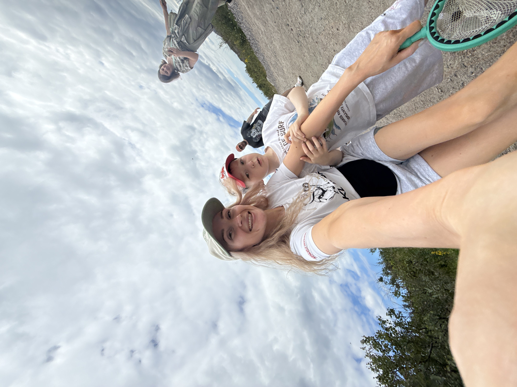 In Teriberka, Russia, a group of people are captured in a selfie outdoors. A woman in the foreground, wearing a white t-shirt and a light green baseball cap, smiles at the camera. Beside her, a child wearing a red and white baseball cap and a white t-shirt is partially visible, with their arm around the woman. Another child in a white t-shirt stands behind them. Further in the background, a man in a black t-shirt and a woman in a patterned top and camouflage pants are walking. The setting appears to be a dirt path with sparse vegetation and trees on either side, under a cloudy sky. The woman in the foreground is holding a green butterfly net.