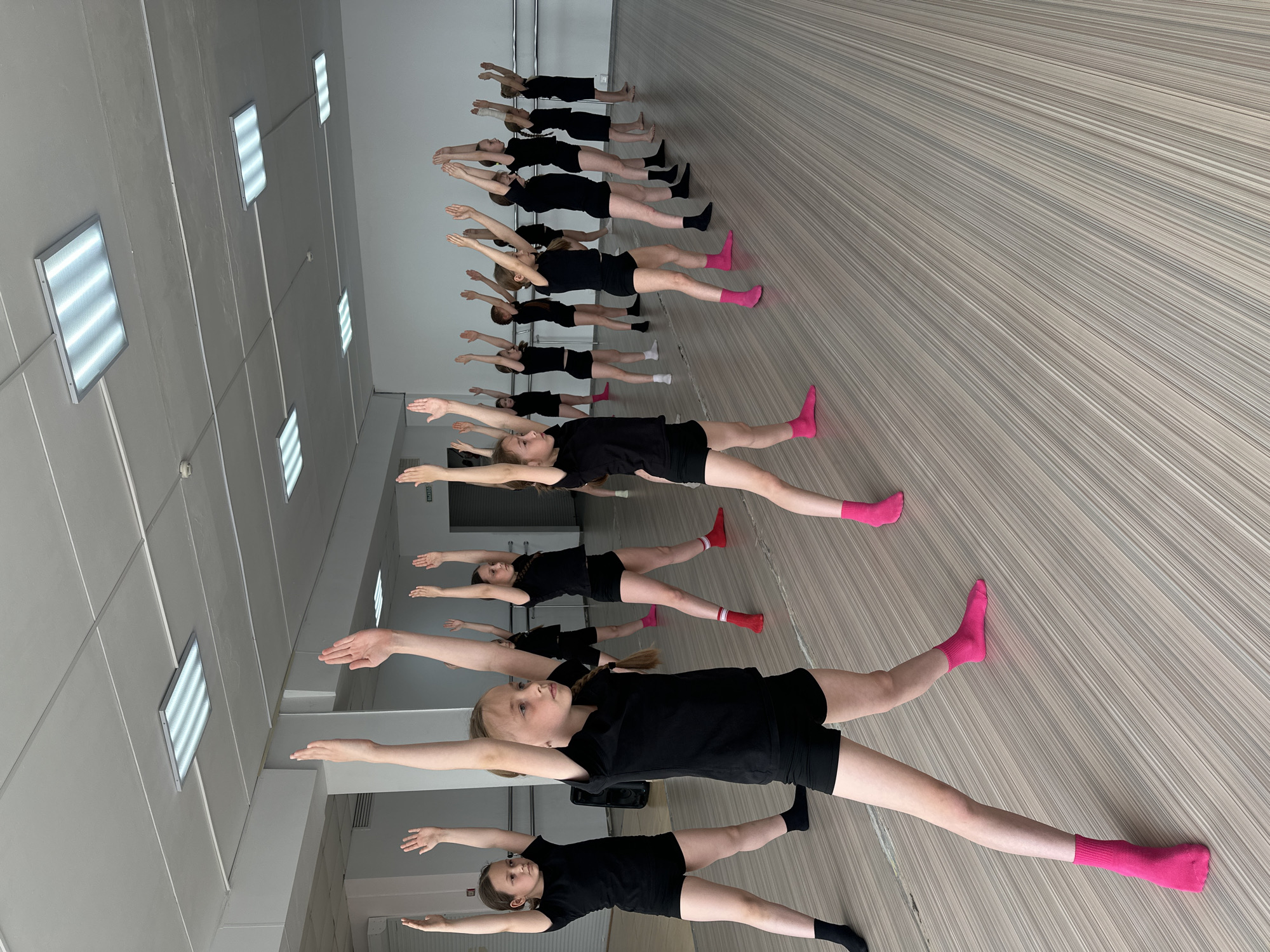 A group of approximately twelve young female dancers are practicing a floor exercise in a dance studio. They are lined up parallel to each other, lying on their backs with legs spread and arms extended horizontally to the sides. They are uniformly dressed in black tops and shorts, with varying colors of socks. The studio is well-lit with rectangular fluorescent lights on the ceiling. The floor is a wood-grain pattern. The location may be Votkinsk, Russia.