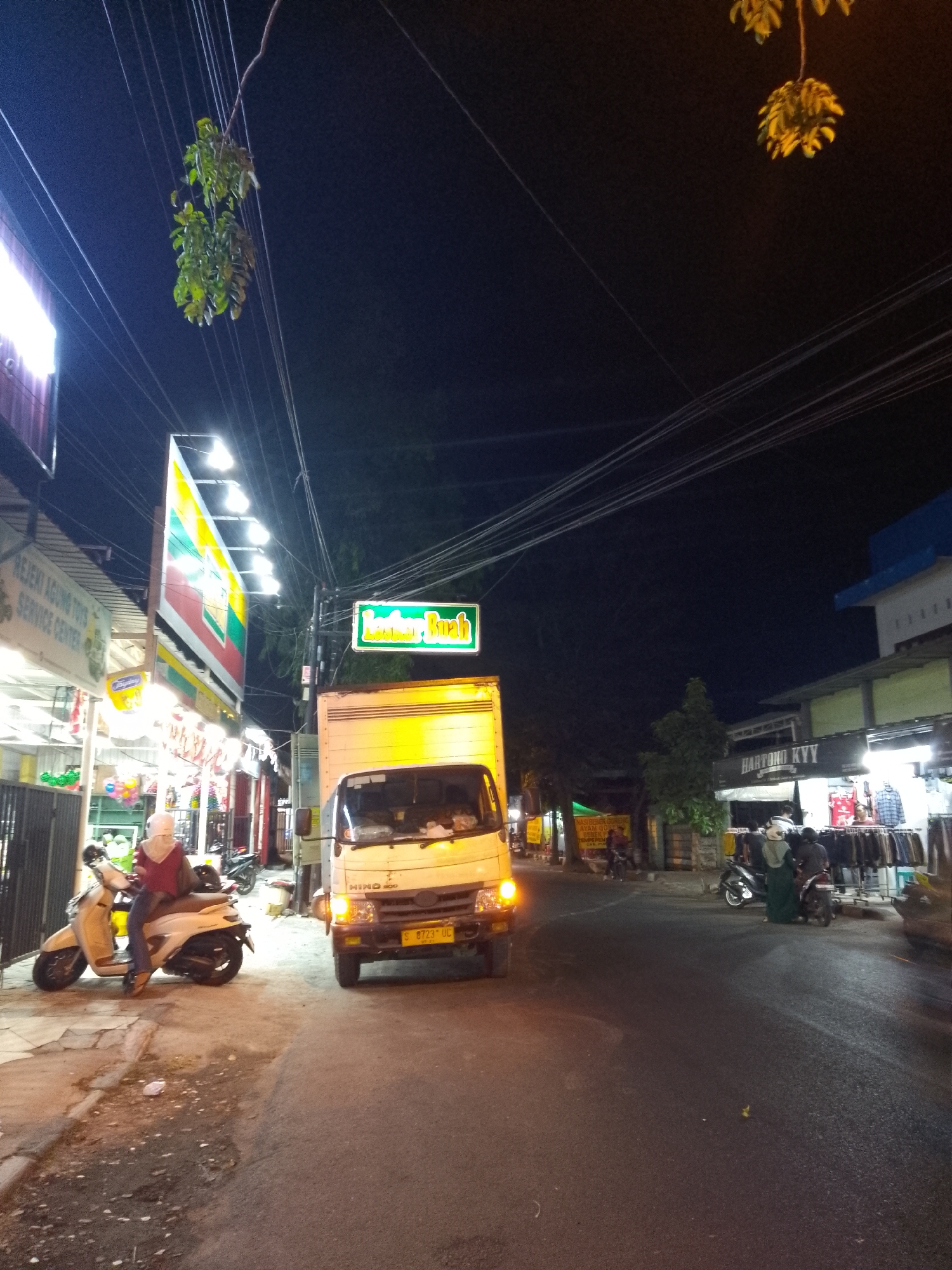 The image depicts a nighttime street scene in Tumenggungan, Indonesia. In the foreground, a yellow truck with license plate "S 9723 UC" is parked on the right side of the road. To the left, a person wearing a hijab and a maroon top is seated on a white scooter. Behind the scooter, a building with bright lights and signage is visible, with text that appears to include "REJEKI AGUNG TOYS SERVICE CENTER". Further down the street, a green illuminated sign reading "LALULALU BUAH" is mounted on a pole. A row of shops lines the right side of the street, displaying clothing on racks. Several other motorcycles are parked in front of these shops, and a few people are browsing. Overhead, numerous power lines crisscross the sky, and clusters of green and yellow foliage hang from branches, illuminated by the streetlights. The overall environment is consistent with a busy commercial street at night.