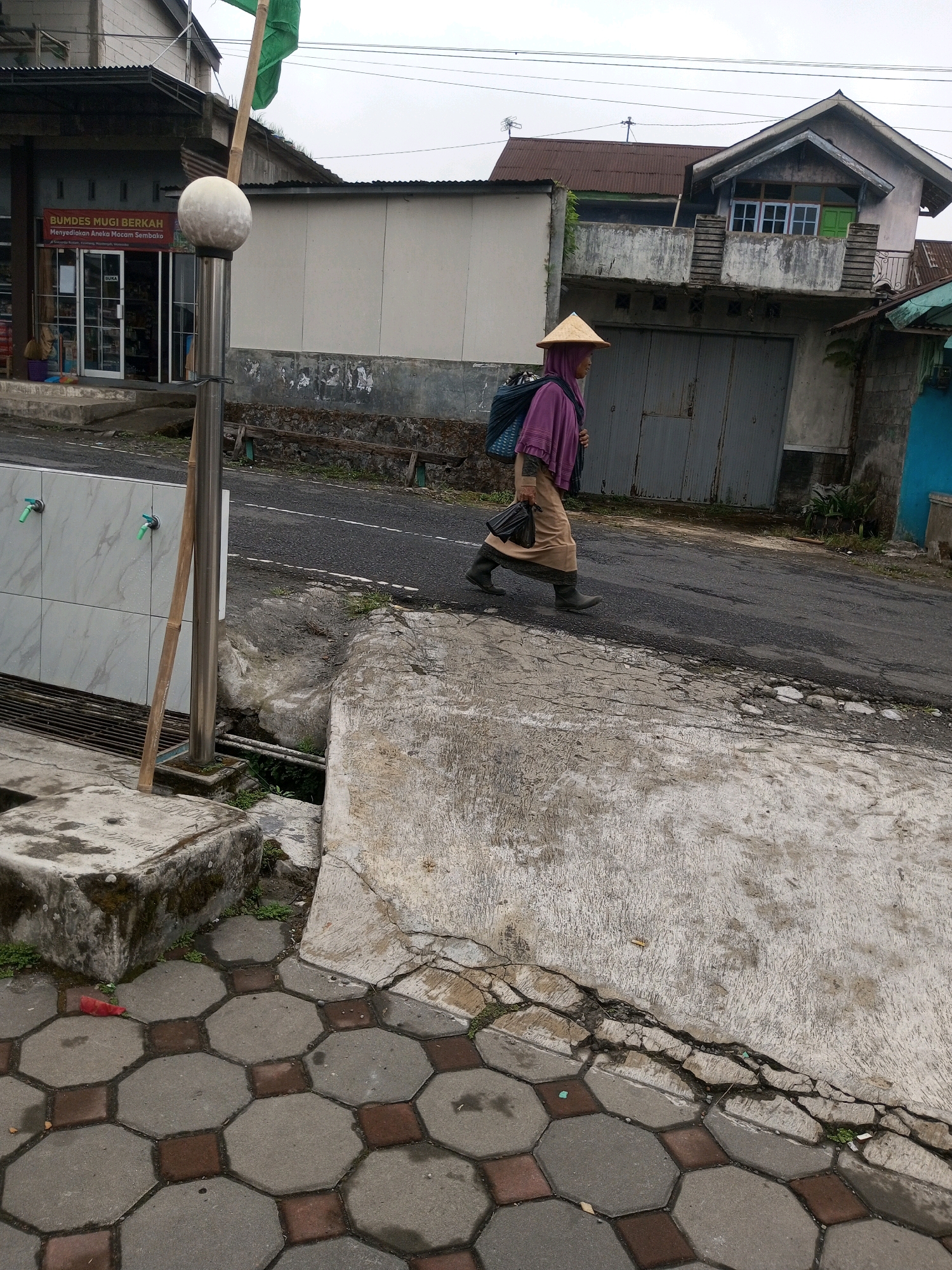 In Keseneng, Indonesia, a person wearing a conical straw hat and a purple headscarf walks along a cracked asphalt road. They are carrying a dark bag in their right hand and a large woven basket on their back. Their clothing consists of a long, beige skirt and dark boots. The person is walking past a weathered building with a gray metal garage door and a second story with a balcony. To the left of the road, a white tiled wall with two green faucets is visible, as well as a metal pole with a spherical finial and a green flag. The foreground features octagonal paving stones. The background shows more buildings and a cloudy sky.