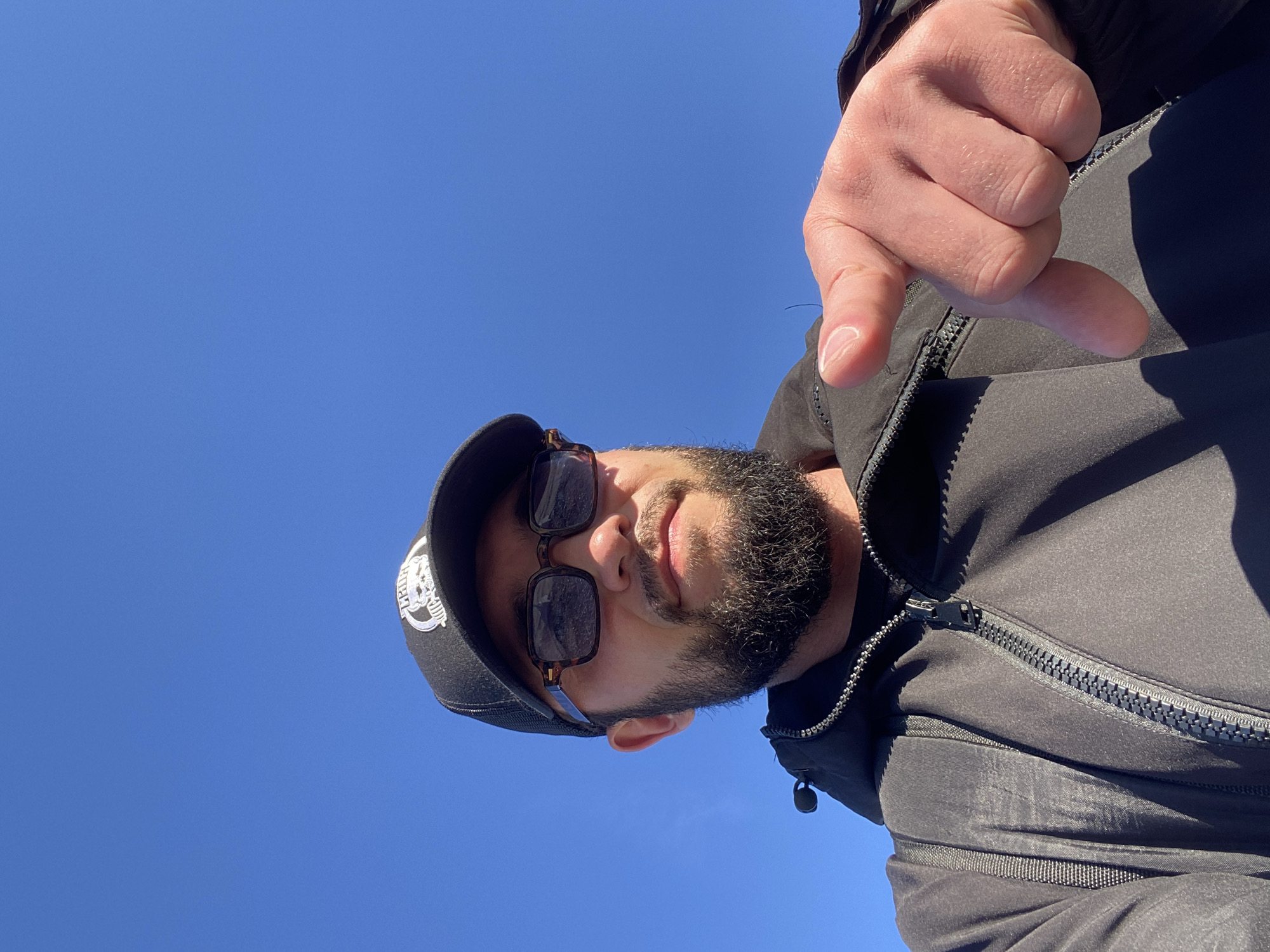 A man is taking a selfie outdoors. He is wearing a black baseball cap, sunglasses, and a black jacket. He has a beard and is pointing his right index finger towards the camera. The background is a clear blue sky. The man is likely outdoors in a non-urban environment, given the lack of city structures and the clear blue sky. The location of Koskolovo, Russia is not ascertainable from the image itself.