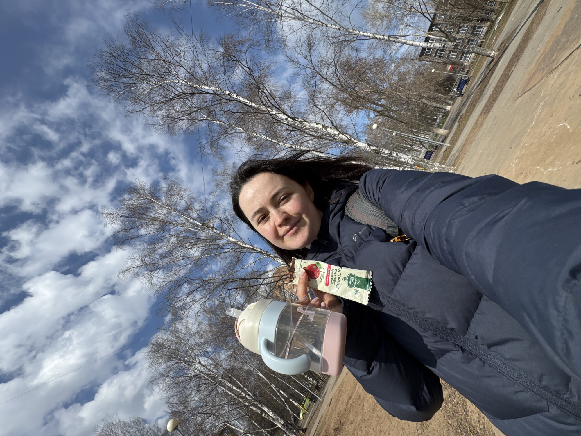 A woman captures a selfie outdoors in what appears to be a park or urban green space in Izhevsk, Russia, on a clear, sunny day. The bare trees, characteristic of birch, suggest late winter or early spring weather.
The woman, with dark hair, smiles faintly at the camera, wearing a dark blue puffer jacket and a visible backpack strap on her right shoulder. She holds two items in her right hand: a transparent plastic water bottle with a light pink base, a light blue handle, and a straw, and a white and green snack bar or pouch. The snack bar displays text in Russian including "БАТОНЧИК" (bar), "ФРУКТОВО-ТВОРОЖНЫЙ" (fruit-curd), and "ЗДОРОВА ЗБС" (likely a health-related brand name, possibly translating to "Healthy ZBS"). A small image of a red fruit, resembling a raspberry or cranberry, is also visible on the packaging.
In the background, tall, bare trees with white trunks line a dirt or gravel path. A multi-story building with numerous windows is visible further back, and street lights dot the landscape. The bright blue sky with scattered white clouds fills the upper half of the frame, indicating pleasant but potentially cool weather conditions. The overall mood conveyed is one of casual outdoor activity and contentment.