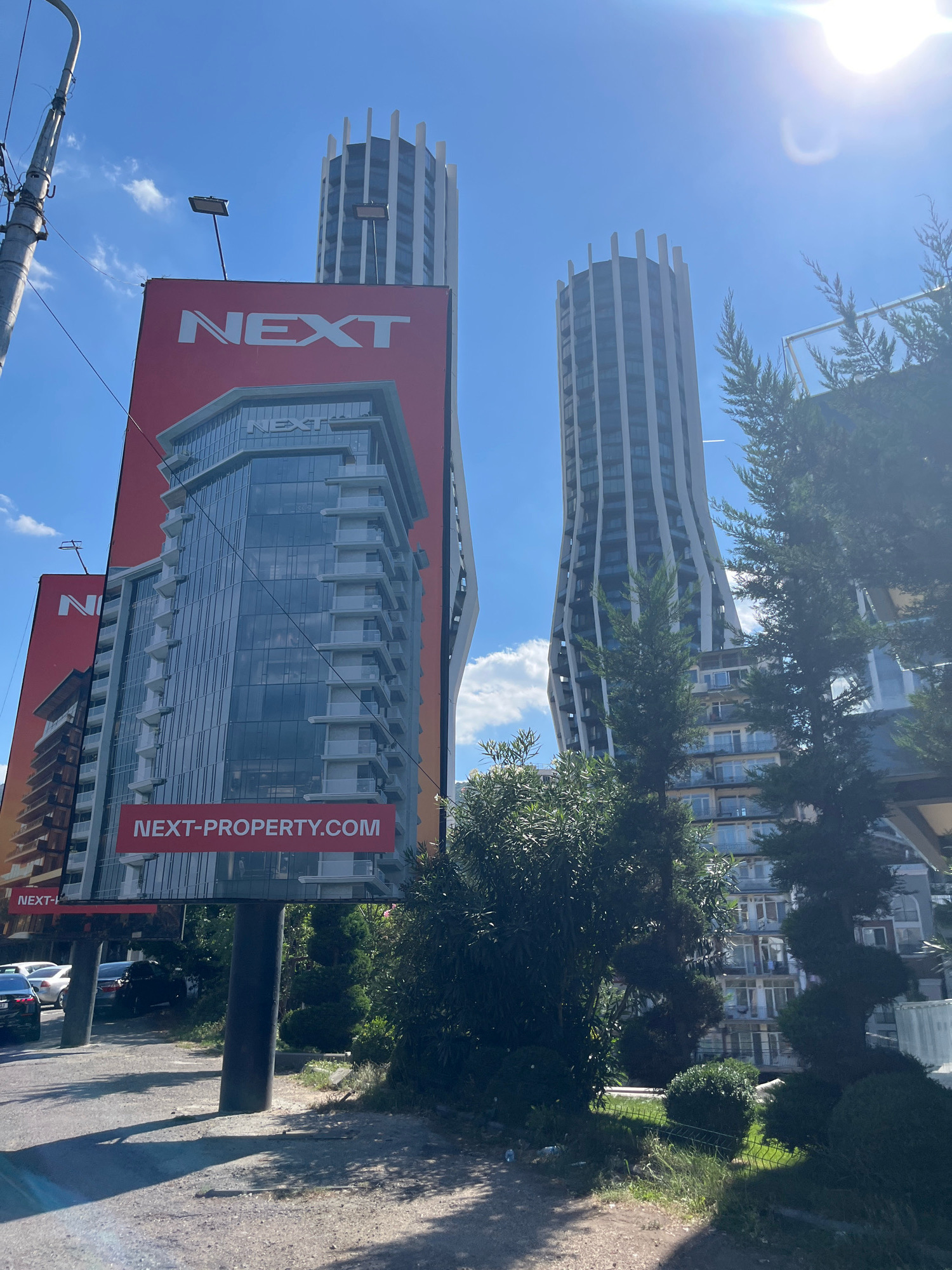 The image shows a billboard advertising "NEXT-PROPERTY.COM" in Tbilisi, Georgia. The billboard features a rendering of a modern building and the text "NEXT." In the background, two contemporary skyscrapers are visible against a clear blue sky. One skyscraper has a distinctive curved facade, while the other is more traditionally rectangular. The scene appears to be a street-level view with some trees and vegetation in the foreground. Cars are parked on the left side of the image. The overall impression is of a developing urban area in Tbilisi.