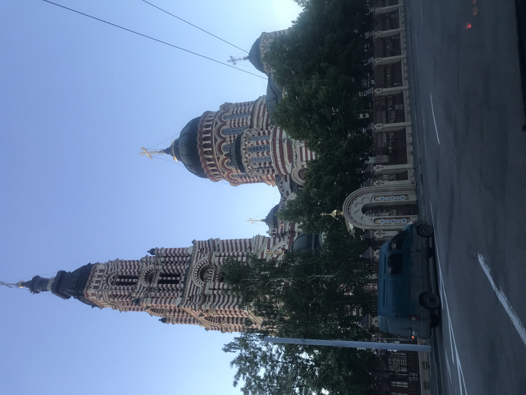 The image shows St. Nicholas Cathedral in Kyiv, Ukraine. The building is a tall, ornate structure with red and white stripes, capped with a black dome and a golden cross. A similar, but smaller building is connected to the first. Lush green trees partially obscure the base of the building. A small blue van is parked on a gray asphalt road. A white line marks the edge of the road. A fence of dark metal poles runs along the bottom edge of the image, behind the blue van. The sky is a clear, light blue. Overhead power lines are visible in the upper part of the frame.