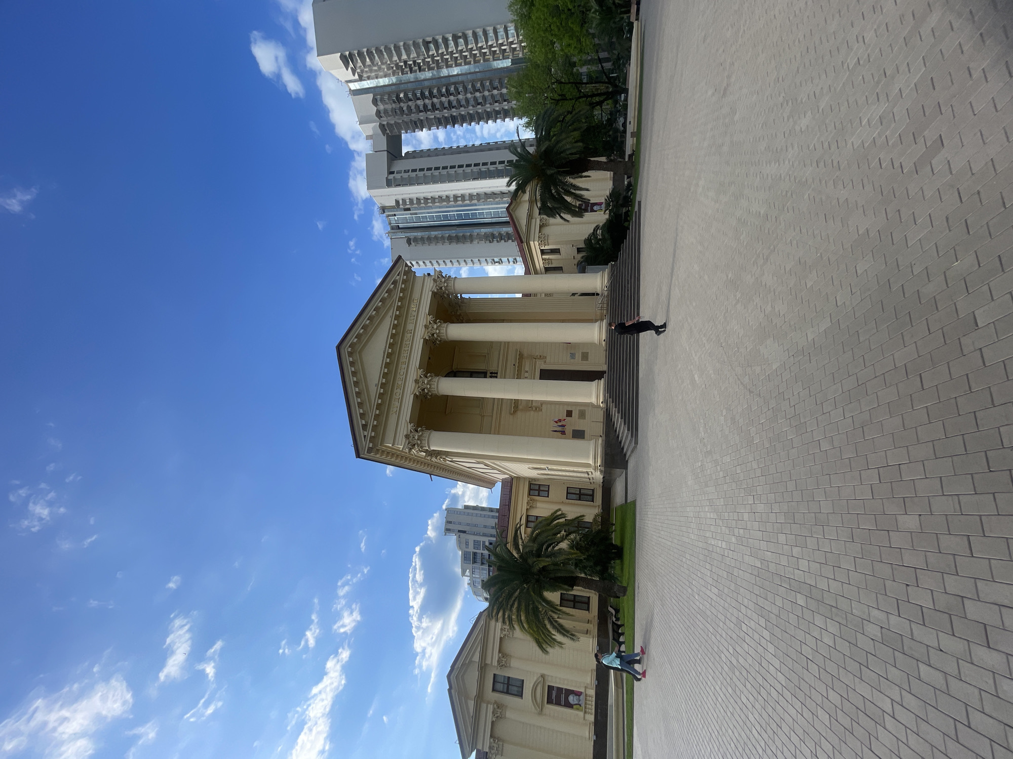 The image depicts a building with classical architecture, featuring large columns and a pediment, juxtaposed with modern high-rise buildings in the background. A person is walking on the stairs leading to the building. Palm trees are visible near the building. The foreground shows a brick-paved area. The sky is blue with scattered clouds. The location is the Sochi Art Museum in Sochi, Russia.