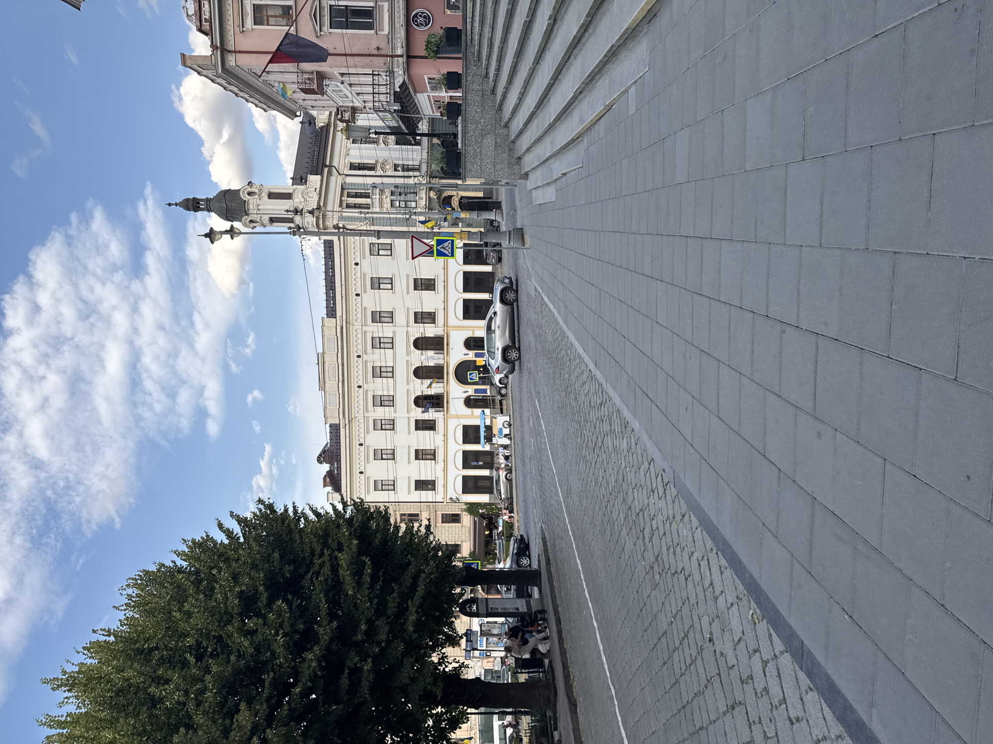 The image depicts a view of a city square with cobblestone and paved sidewalks. In the foreground, a wide paved walkway extends into the distance. To the left, a large, leafy green tree stands prominently. Behind the tree, a street is visible with parked cars and a few people seated on benches. Further back, several buildings line the square. One building on the right features a prominent tower with a dome and a clock. Flags are visible on some of the buildings. A set of stone stairs leads up to one of the buildings on the right. Several cars are driving or parked on the street. The sky is blue with scattered white clouds. The scene appears to be in Chernivtsi, Ukraine, likely a central square or pedestrian area.