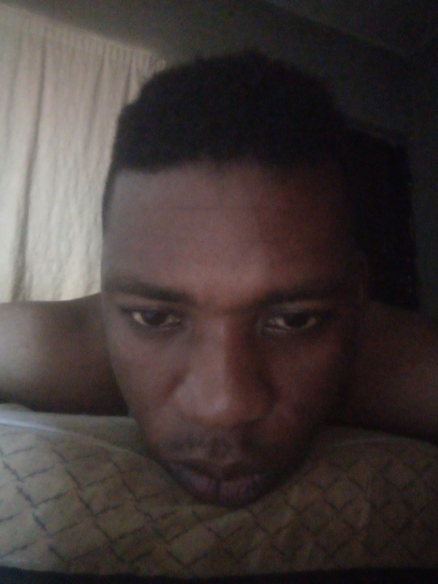 A close-up, low-angle shot of a man with dark hair and skin, lying down. He is resting on a textured, quilted surface, possibly a pillow or comforter. His shoulders are visible in the frame. Behind him, there is a light-colored curtain. The location is unknown.