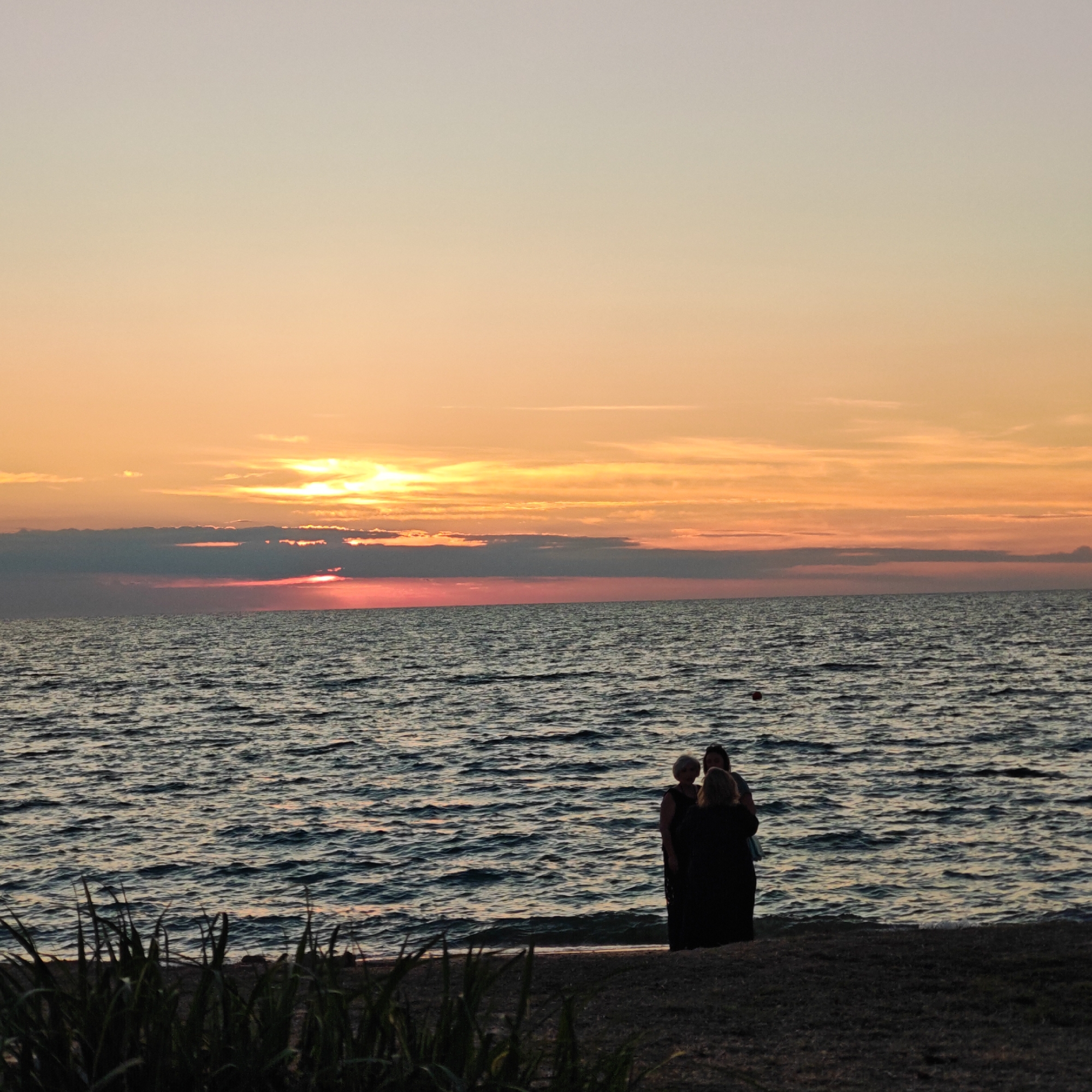A sunset casts an orange and pink glow across the sky over a body of water. The water is dark and choppy with visible waves. On the shore in the foreground, silhouetted figures of three people are gathered. The beach appears to be sandy and dark, with some green vegetation visible in the lower left corner of the image.
