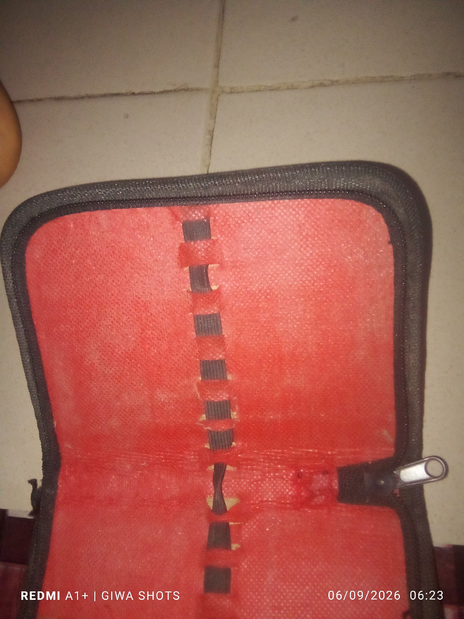 An opened red case with black trim and a zipper is laid on a tiled floor. The interior of the case has multiple dark grey elastic straps running vertically down the center to hold items. A timestamp in the bottom right corner of the image reads "06/09/2026 06:23." Text at the bottom left corner reads "REDMI A1+| GIWA SHOTS."