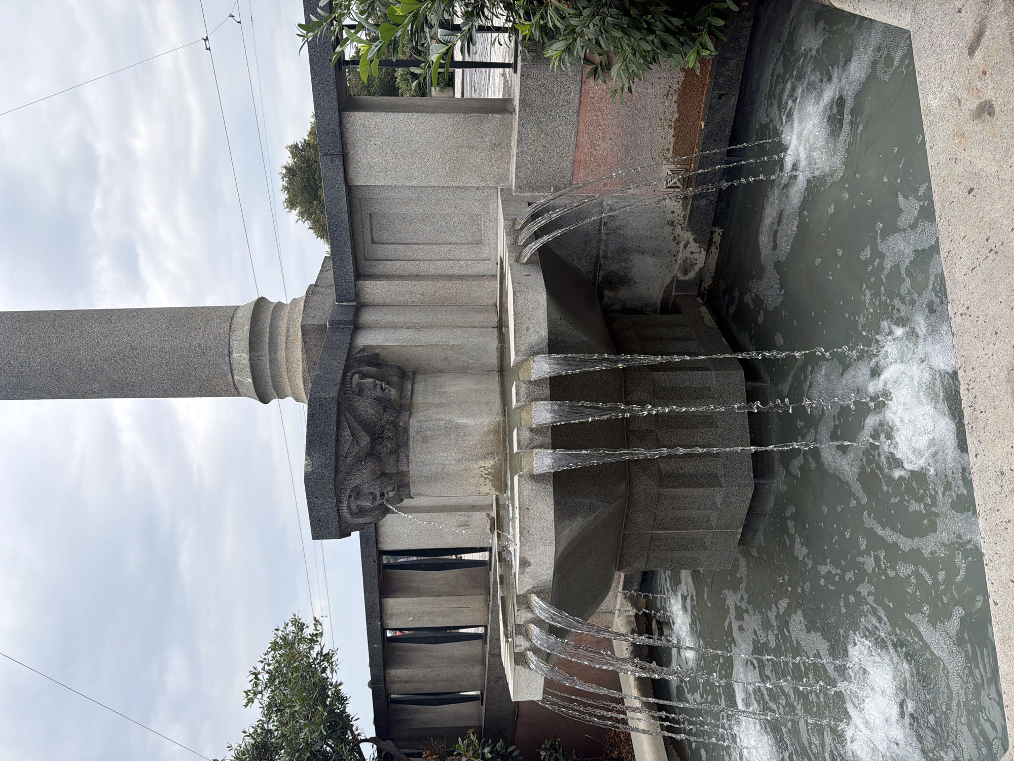 This image depicts a stone fountain with water cascading from multiple spouts into a basin. The fountain is adorned with carved stone faces. In the background, there is a large stone pillar, a railing, and greenery. Overhead, there are power lines against a cloudy sky. The scene appears to be outdoors, possibly in a public space. The style of the fountain and surrounding architecture suggests it could be located in Prague, Czechia, as it resembles other fountains found in the city.