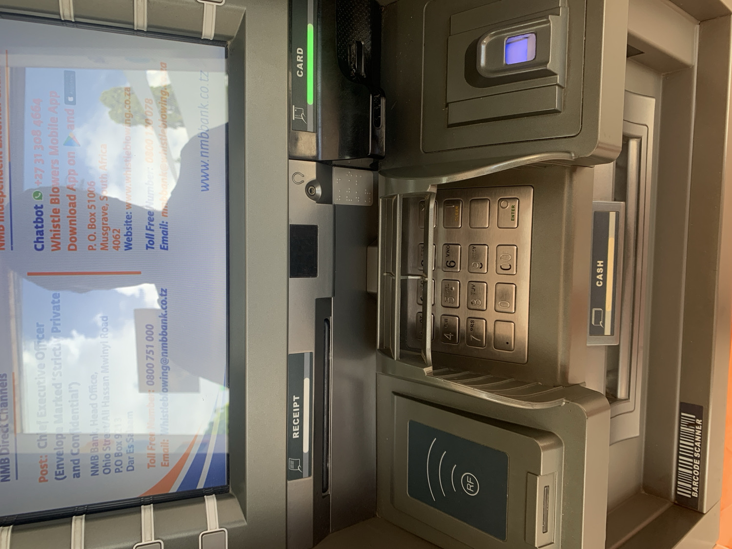 This close-up shot captures a modern automated teller machine (ATM) of NMB Bank, likely situated indoors or under cover in Dar es-Salaam, Tanzania, given the on-screen information. The ATM appears to be in an idle state, displaying contact information and service details on its screen.
The screen, occupying the left side of the frame, shows various "NMB Direct Channels." It lists contact information for the "Chief Executive Officer" with an address for "NMB Bank, Head Office, Ohio Street/Ali Hassan Mwinyi Road, P.O Box 9213, Dar Es Salaam," confirming the Tanzanian context. A Tanzanian toll-free number (0800 751 000) and an email address for whistleblowing (Whistleblowing@nmbbank.co.tz) are also visible, along with the website "www.nmbbank.co.tz". Additionally, details for a "Whistle Blowers Mobile App" are displayed, including a South African chatbot number (+27 31 308 4664), a P.O. Box address in Musgrave, South Africa, and a South African whistleblowing website and toll-free number.
To the right of the screen are the ATM's functional components. A "CARD" slot, indicated by a green light, is at the top right. Below it is the numeric keypad with "CLEAR," "ENTER," and "CANCEL" buttons, accompanied by a smaller, blue-lit biometric scanner or indicator above. A slot labeled "CASH" is positioned to the right of the keypad, and further down, a "RECEIPT" slot is visible. At the bottom left, a contactless payment reader with an "(RF)" and antenna symbol is present, while a vertical "BARCODE SCANNER" is noted along the bottom right edge. No individuals are present in the image, implying the ATM is ready for use.