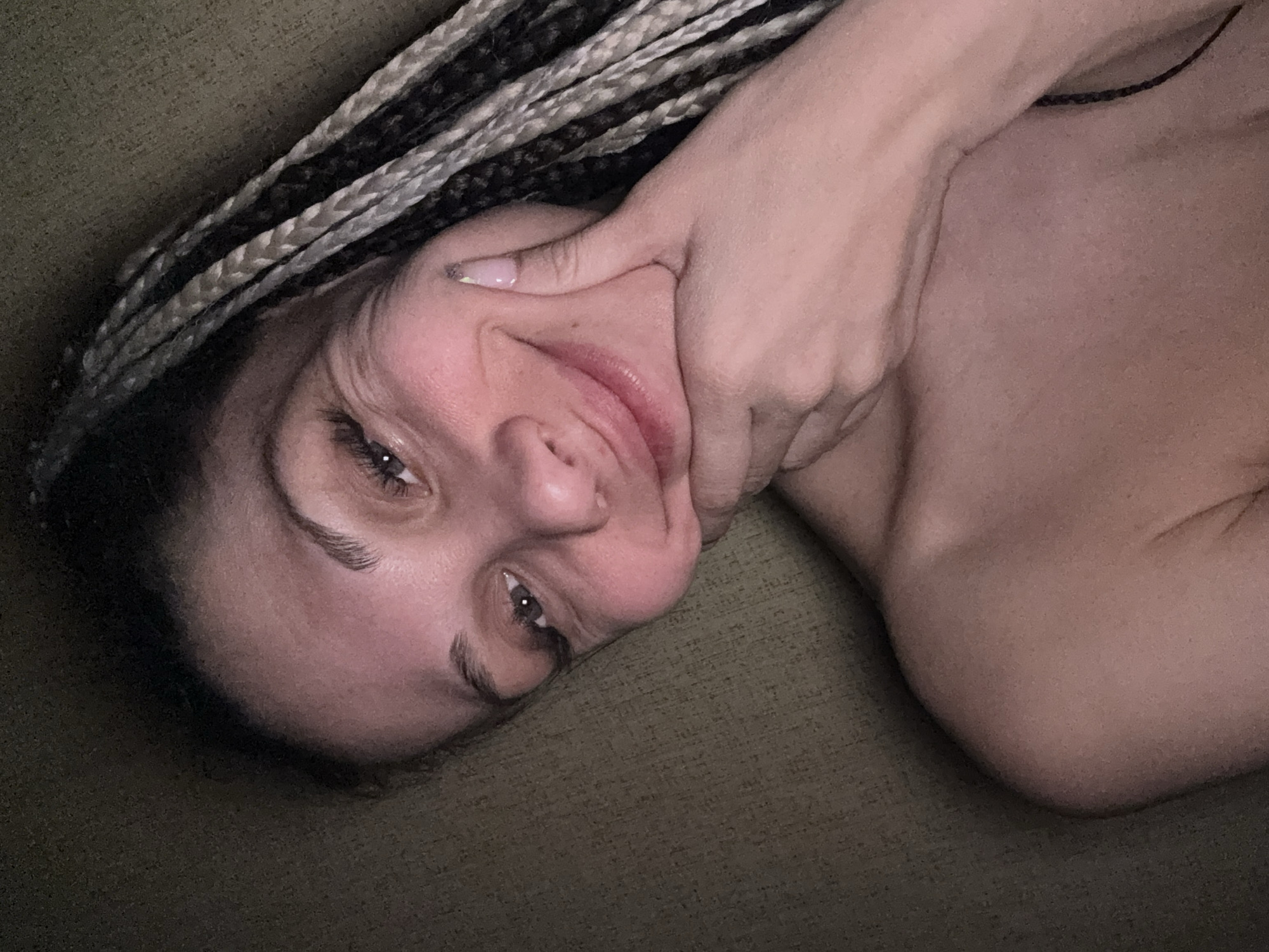 The image shows a close-up, angled shot of a woman lying down in an indoor setting. Her head is tilted, and she is looking directly at the viewer with open eyes and a slight smile. She has dark hair styled in box braids with prominent lighter, possibly blonde or grey, accents. Her right hand is playfully placed on her face, seemingly pulling her cheek or jawline, which creates a slightly distorted but lighthearted expression. Her bare shoulder and upper chest are visible, suggesting she is partially unclothed. The background consists of a dark, textured fabric, likely upholstery or bedding, indicating a relaxed and intimate indoor environment. The lighting is subdued, suggesting evening or nighttime. No text is visible in the image. The overall impression is one of casual playfulness.