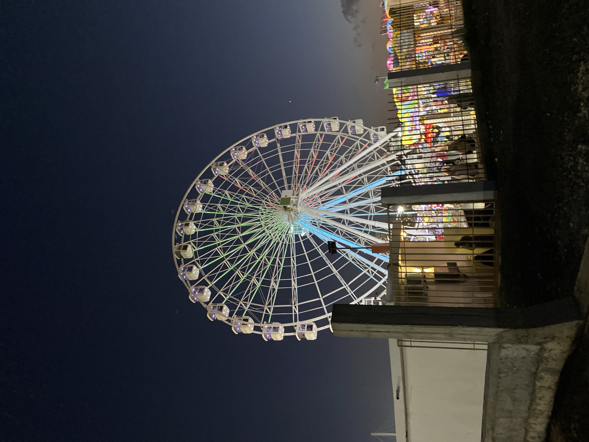 An illuminated Ferris wheel is the central subject of this nighttime outdoor scene, likely located at a fairground or amusement park in Hussein Dey, Algeria. The Ferris wheel is white with multi-colored lights, primarily green, blue, and red, outlining its structure and cabins. It dominates the upper half of the image against a dark, clear night sky. In the foreground, a concrete wall and a metal fence enclose a pathway and some structures. Behind the fence, in the midground, are various carnival rides and attractions, also illuminated with colorful lights, though less distinctly visible due to distance and the fence. A few indistinct figures can be seen through the fence, appearing to be patrons or staff. The ground in the immediate foreground is dark and appears to be unpaved. The overall atmosphere suggests a lively evening at an entertainment venue.