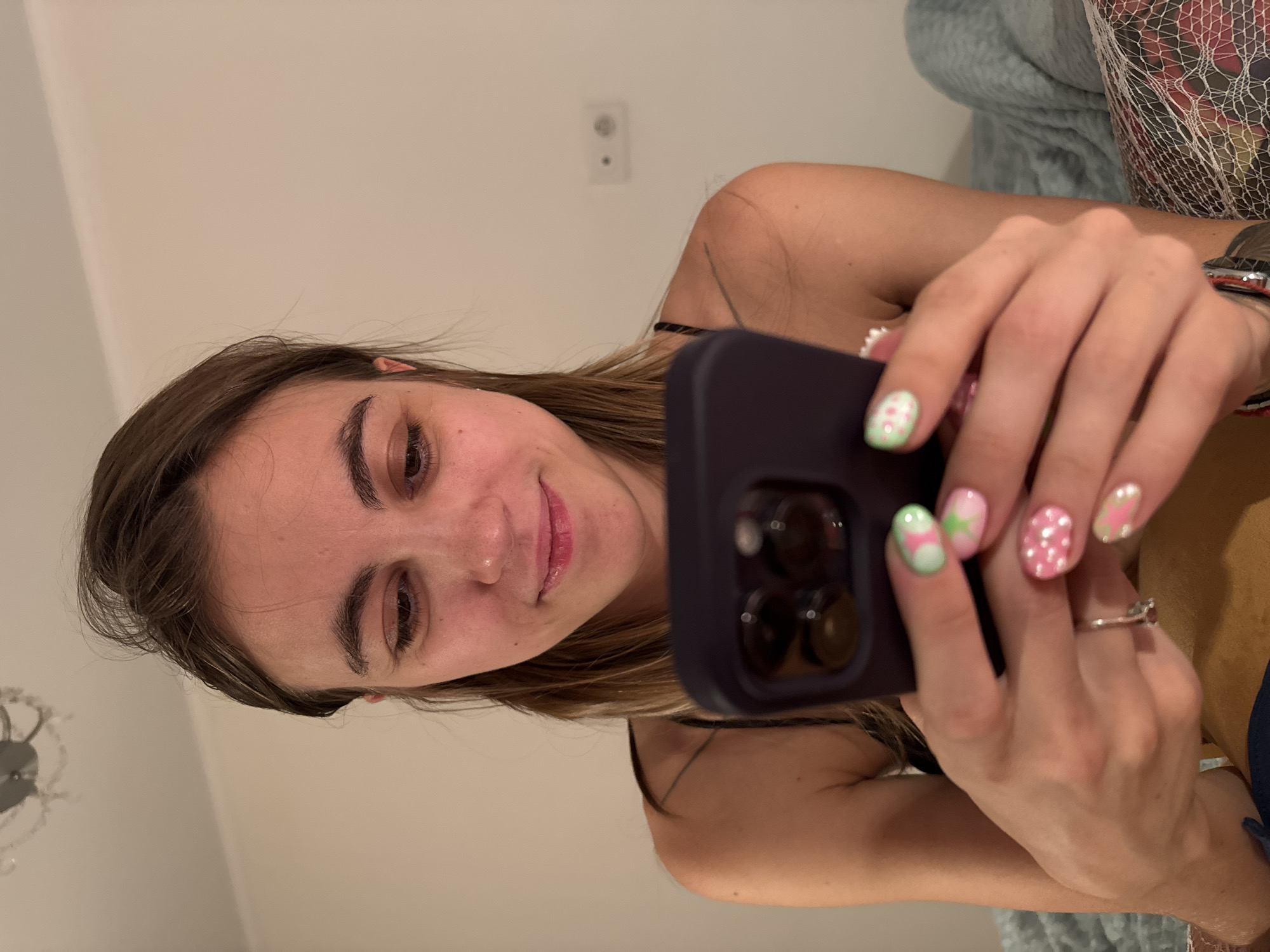 A young woman with brown hair is looking down at her phone, which she holds in her hands. She is wearing a black spaghetti strap top. Her nails are painted with a colorful design, featuring pink and green with white polka dots and floral accents. She has a subtle smile on her face. The background is a light-colored wall with a power outlet visible. There is a ceiling fixture and a light-colored blanket to the right. The scene appears to be indoors, possibly a bedroom. There is no information in the image to indicate the location is Moscow, Russia.