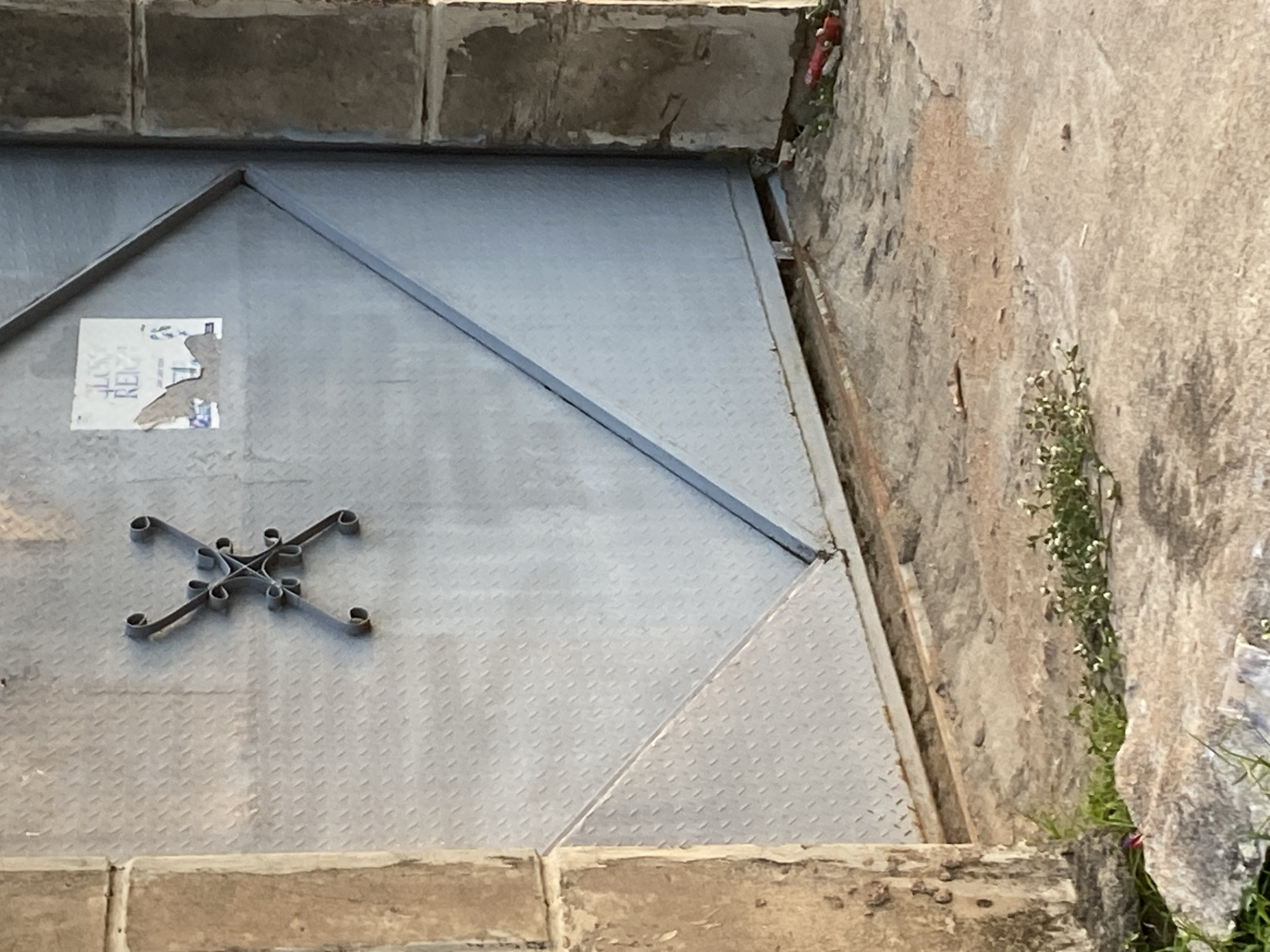 The image shows a metal gate with a decorative metal cross design in the center. There is a torn poster on the upper right side of the gate. To the left and right of the gate are concrete pillars. The ground in front of the gate is concrete with patches of dirt and some small green plants. The scene appears to be an exterior shot in Jauro Yinu, Nigeria.