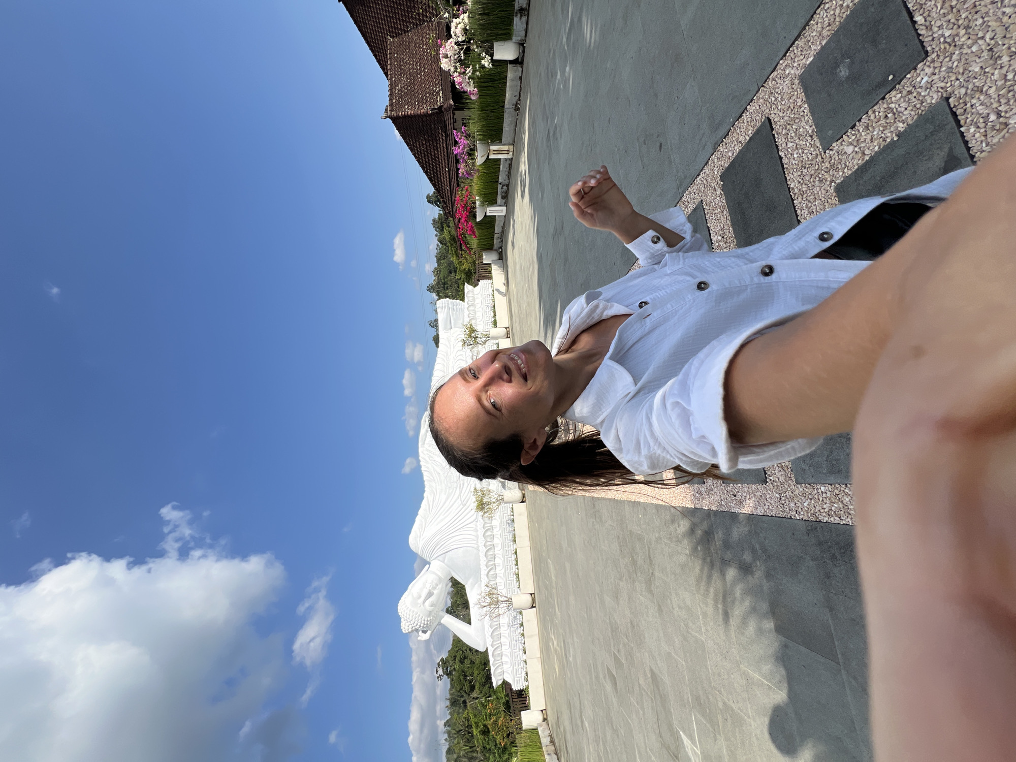 A woman, appearing to be in her late 20s or early 30s, is taking a selfie outdoors in front of a large white reclining Buddha statue. She has long, dark brown hair pulled back in a ponytail and is wearing a white, button-up shirt. She is smiling at the camera and her right hand is partially visible, reaching towards it.
The Buddha statue is enormous and is positioned on a raised platform. Behind the statue, there are lush green trees and a blue sky with scattered white clouds. To the right of the statue, there is a building with a reddish-brown tile roof and a row of flowering plants in white planters. The ground in the foreground is paved with large gray tiles and interspersed with gravel pathways. The scene appears to be a serene outdoor temple or garden complex. The location is identified as the Pupuan Buddha Temple in Pupuan, Bali, Indonesia.