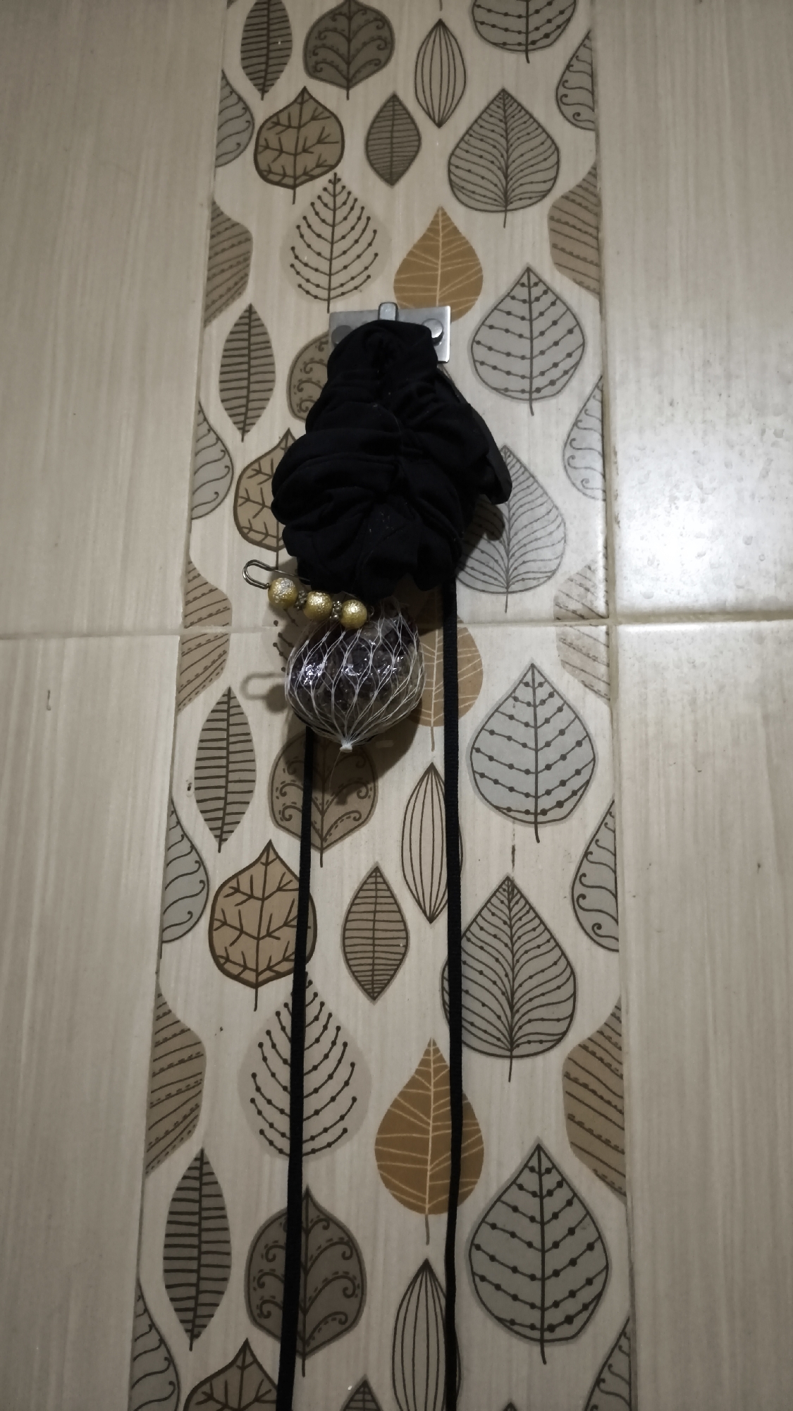 A close-up, eye-level shot shows a black scrunchie and a dangling decorative ball attached to a silver hook mounted on a decorative vertical panel. The panel features a repeating pattern of various stylized leaves in shades of brown, gray, and beige against a white background. Two black strings hang vertically from the scrunchie, with the decorative ball suspended from one of them. The ball is enclosed in a transparent mesh-like material, revealing a dark interior, and is adorned with small, gold, bead-like accents at the top. The hook is positioned centrally on the decorative panel, which appears to be mounted on a tiled wall, with visible grout lines suggesting a bathroom or kitchen environment. The lighting is somewhat dim, casting subtle shadows. The scene is located in a residential setting, likely in the city of South Jakarta, Indonesia.
