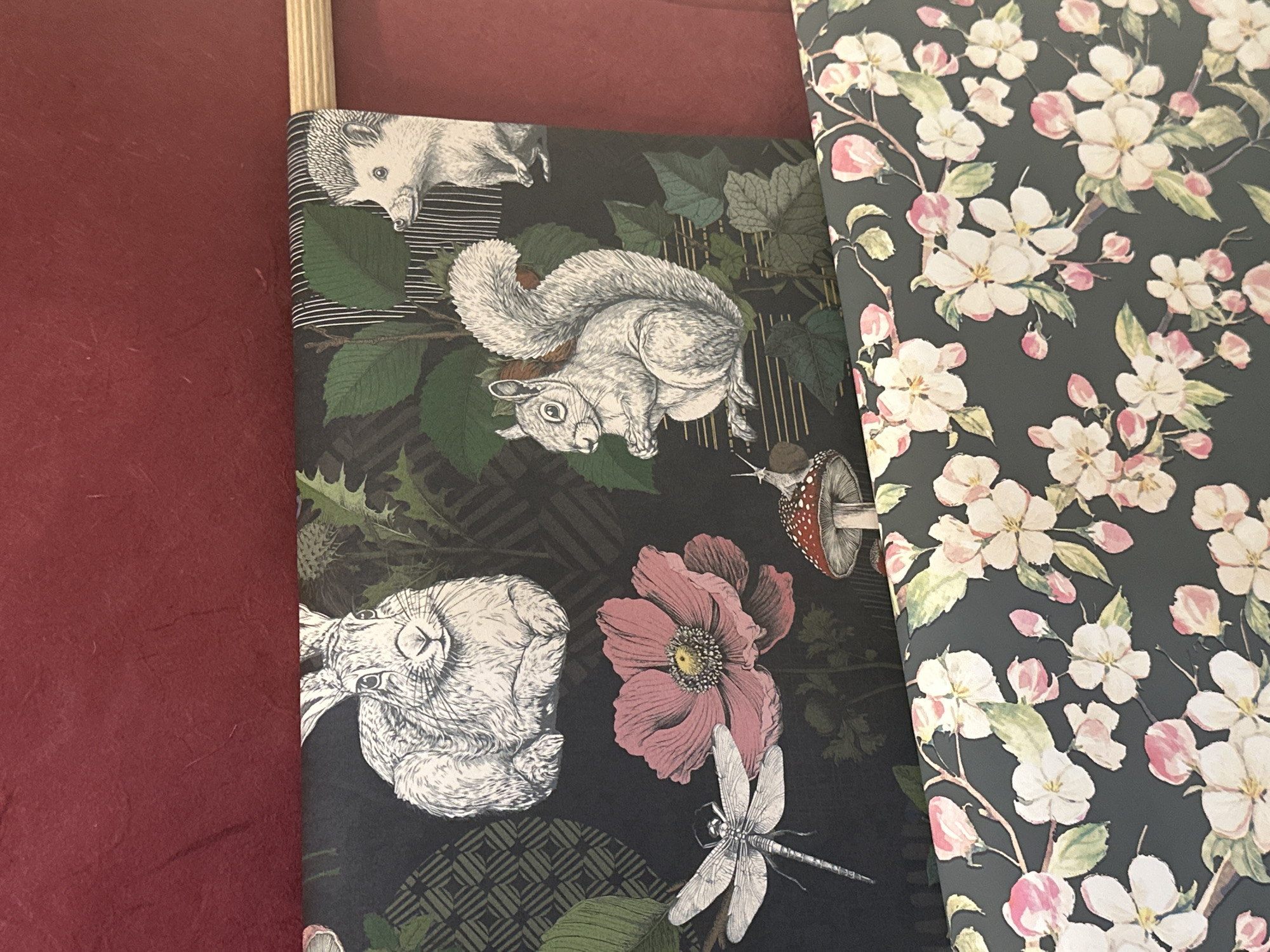 The image shows two rolls of wallpaper. The roll on the left features a dark green background with images of a hedgehog, squirrel, hare, pink flower, dragonfly, and mushroom. The roll on the right has a floral pattern of white and pink blossoms on a dark green background. The setting appears to be indoors. The city is not discernible from the image.