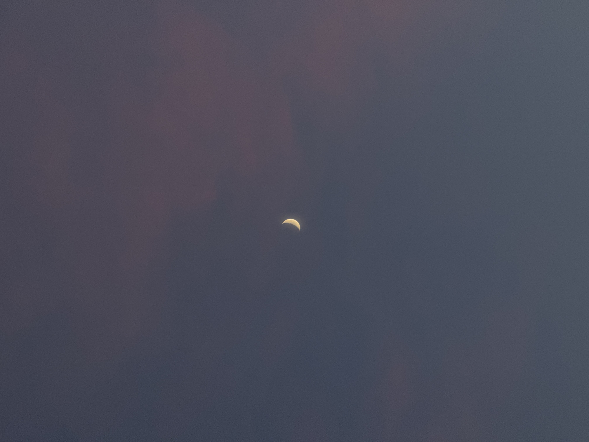 A thin, crescent moon is visible in a dusky sky, slightly to the left of the center of the frame. The sky is a deep, muted blue with hints of purple and pink in the upper right quadrant, suggesting either twilight or the presence of thin clouds. The overall tone of the image is serene and atmospheric, with the moon being the sole distinct celestial object. There are no discernible features related to the city of Mikhaylovsk, Russia, or any other geographical indicators within the image. The image is a minimalist depiction of a night sky.