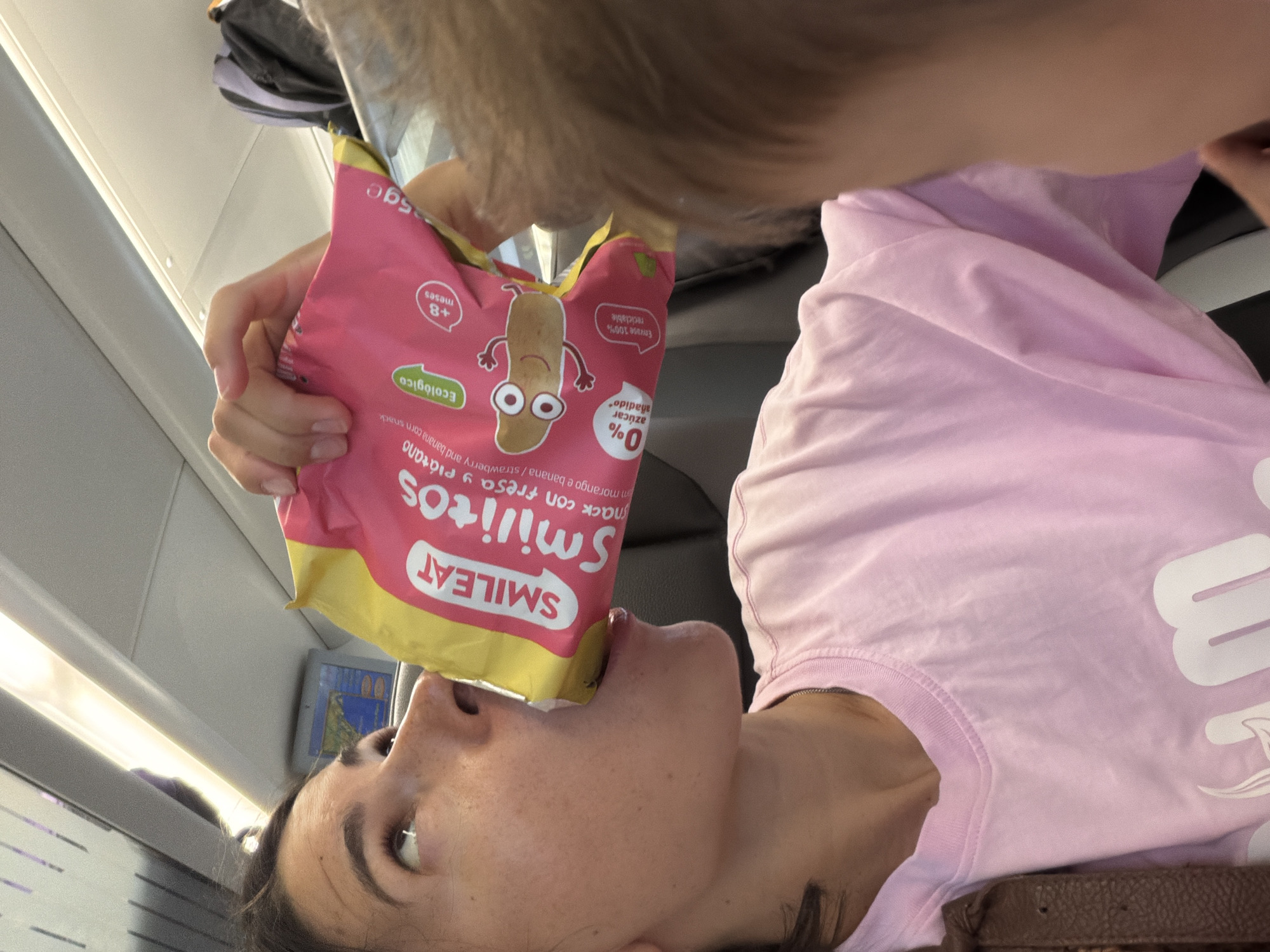 An adult woman wearing a pink t-shirt is shown from the chest up, leaning forward and drinking directly from a pink and yellow bag of SMILEAT "Smilítos" snack. The snack bag features an illustration of a cartoonish brown bean character with eyes and arms, and text in Spanish and English indicating it is a strawberry and banana corn snack with 0% added sugar and is ecological. The background appears to be the interior of a train, with a screen displaying a map visible above and to the left of the woman's head. A portion of a child's head and hair is visible in the foreground on the right side of the image, obscuring part of the woman's face and body. The overall lighting suggests an indoor, daytime environment. There is no indication of the city of Tarragona, Spain in this image.