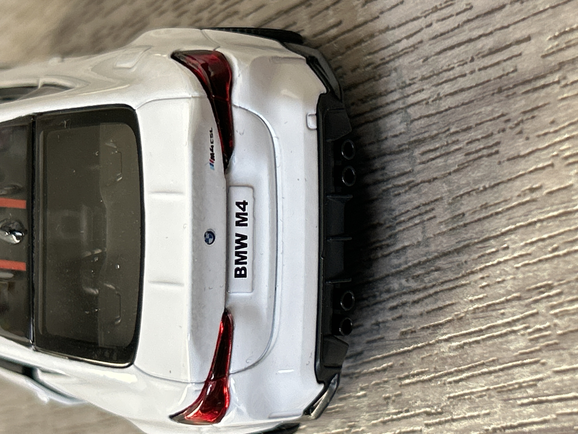 The image shows the rear of a white BMW M4 car. The license plate reads "BMW M4". The car has a distinctive taillight design with red accents. There are four exhaust pipes visible on the right side. The car is resting on a wooden surface. No people are visible. There is no indication that this is in Strasbourg, France.