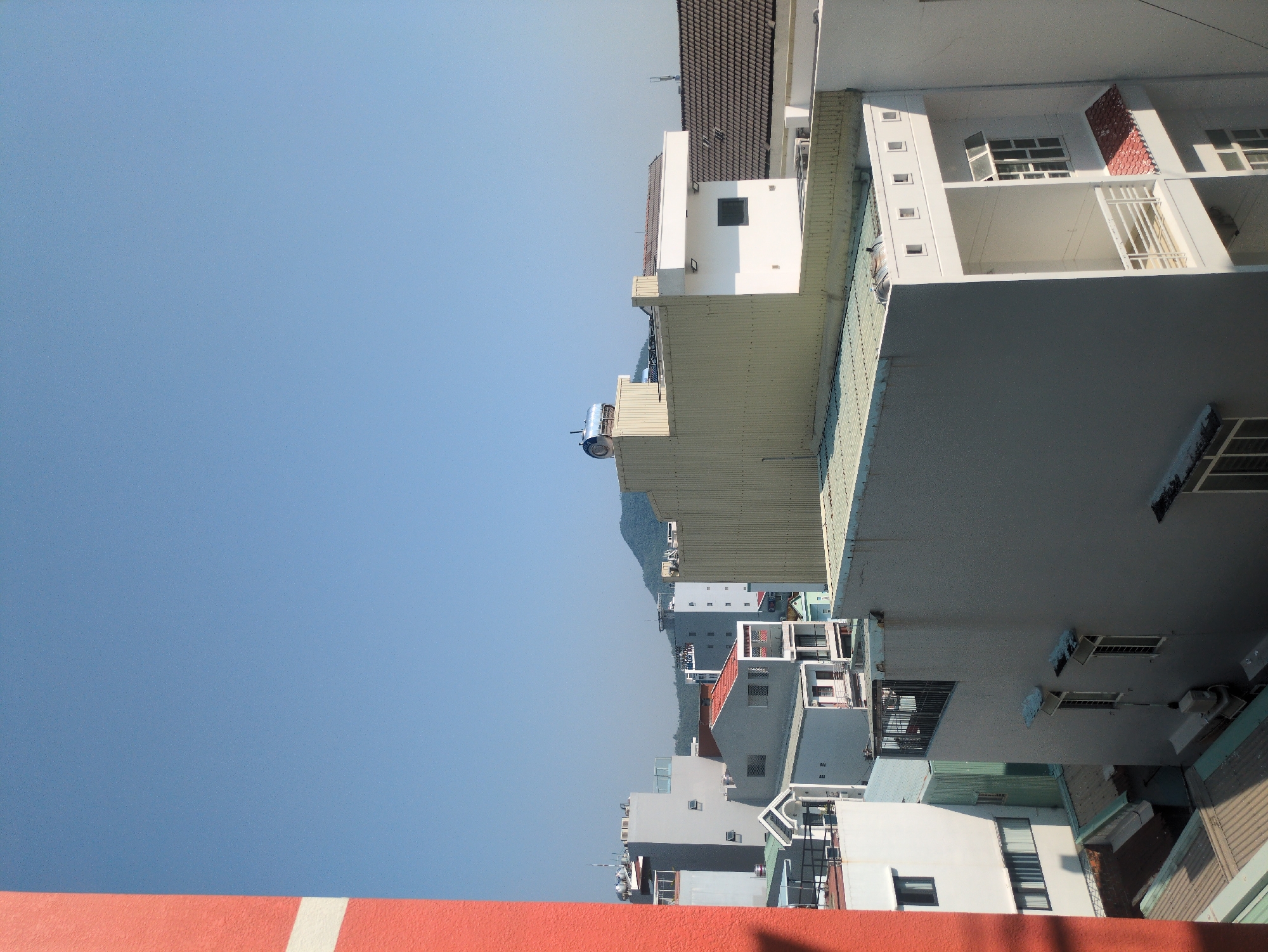 The image shows a view looking up at buildings in South Quy Nhon Ward, Vietnam. The sky is clear and blue. The buildings are a mix of white, gray, and red colors, and vary in height. Some have balconies and windows. A mountain range can be seen in the distance.