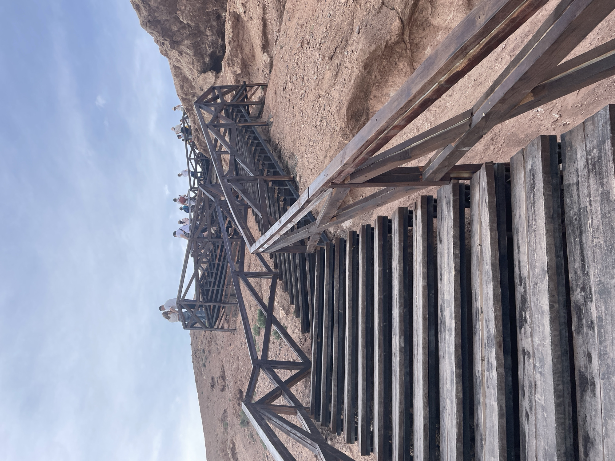 An upward-angle, eye-level shot captures a wooden staircase ascending a red, rocky hillside under a hazy, pale blue sky. The staircase, constructed from dark wooden planks and railings, dominates the foreground and middle ground. Several individuals, dressed in casual, light-colored clothing, are visible on different levels of the staircase and on the hillside above, appearing to be tourists. The rocky terrain features sparse vegetation. The scene depicts a path leading up a natural elevation, possibly a geological formation, with man-made infrastructure for visitor access. The location is the Charyn Canyon in the Tamerlik region of Kazakhstan.
