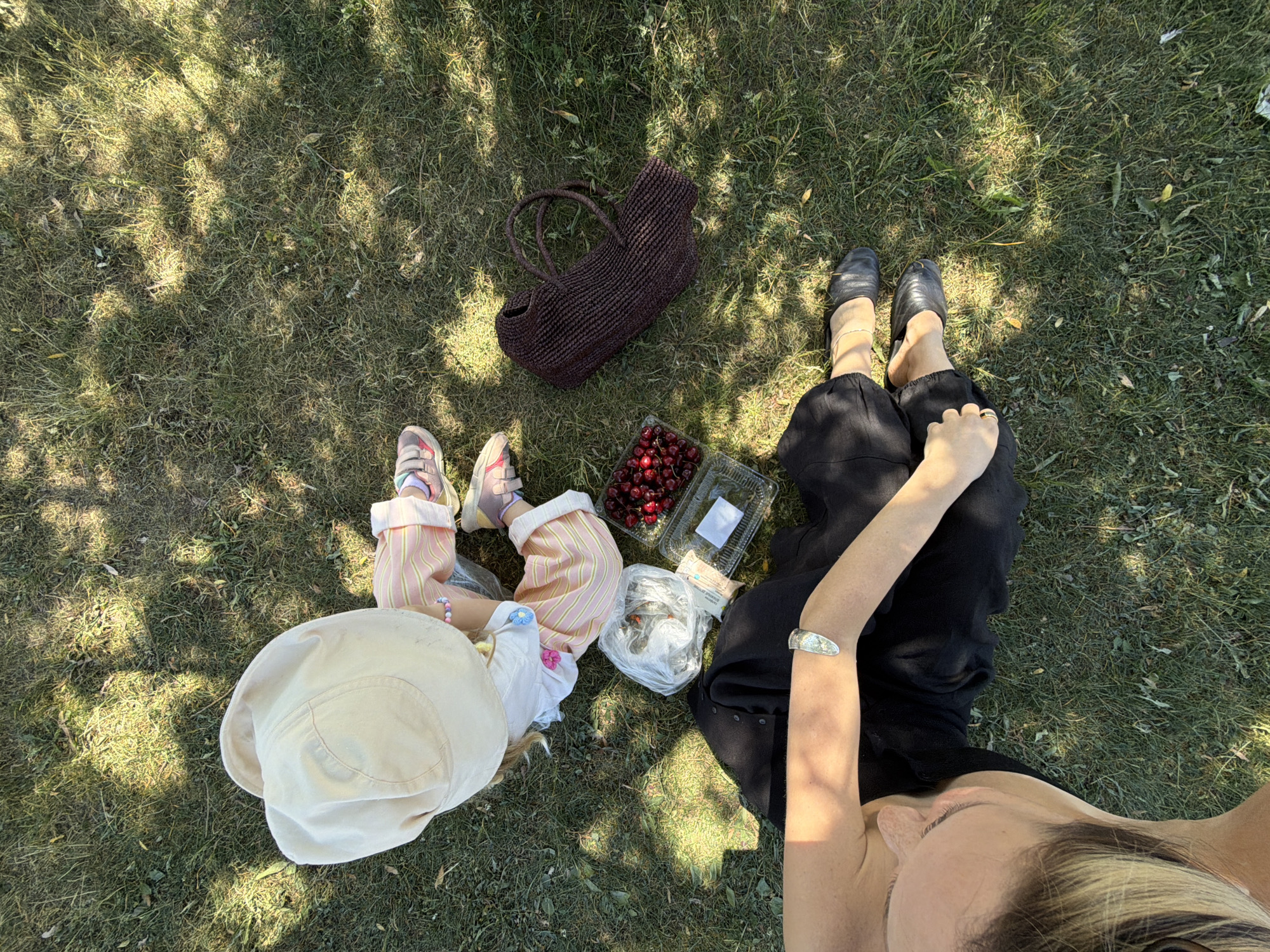 An overhead, sunlit view depicts an outdoor grassy area in the city of Khimki, Russia. Two individuals, an adult and a child, are seated or reclining on the patchy green grass under dappled sunlight.
The adult, partially visible at the bottom-left of the frame, wears a black top, black flowing pants, and black flat shoes. A silver-toned cuff bracelet is on her right wrist, and a ring is on her right hand. Her light-colored hair is visible as her head is turned to the left.
The child, seated at the top-center with their back to the camera, wears a light-colored bucket hat, a white t-shirt with small floral appliqués, light pink and white striped pants, and light-colored sneakers. A beaded bracelet is on their left wrist.
Between the individuals, on the grass, are a clear plastic container of dark red cherries, a clear plastic bag containing white items (likely food), a small wrapped packet, and an empty or partially full clear plastic container. To the right, a dark brown woven bag with handles lies on the grass.
