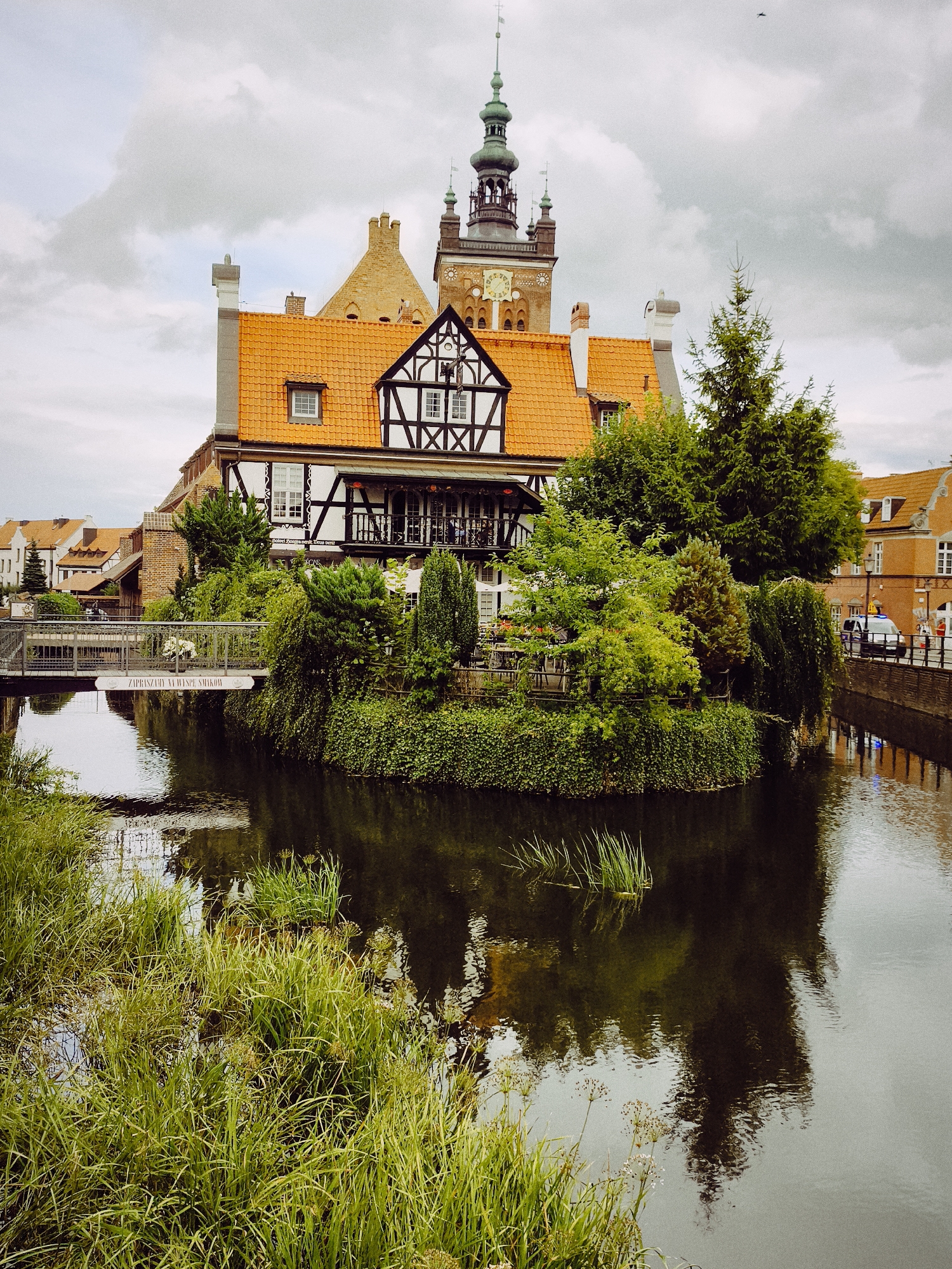 The image captures a view of the Motława River in Gdańsk, Poland, specifically near the historic Crane (Żuraw). In the foreground, tall green grass grows along the bank of the river, with reflections of trees and buildings visible on the water's surface. A footbridge crosses the river, connecting the street on the right bank to the island with the building on the left.
The central subject is a building with traditional Dutch-style architecture, featuring a prominent orange tiled roof and a black and white timber-framed facade. A tall tower, characteristic of Gdańsk's historic architecture, rises behind this building. The tower has a green spire and a clock face. Surrounding the building is lush greenery, including trees and bushes, creating a picturesque island setting. On the right side of the image, a street runs alongside the river, with a parked police car and a few pedestrians visible. The sky is overcast with scattered clouds.