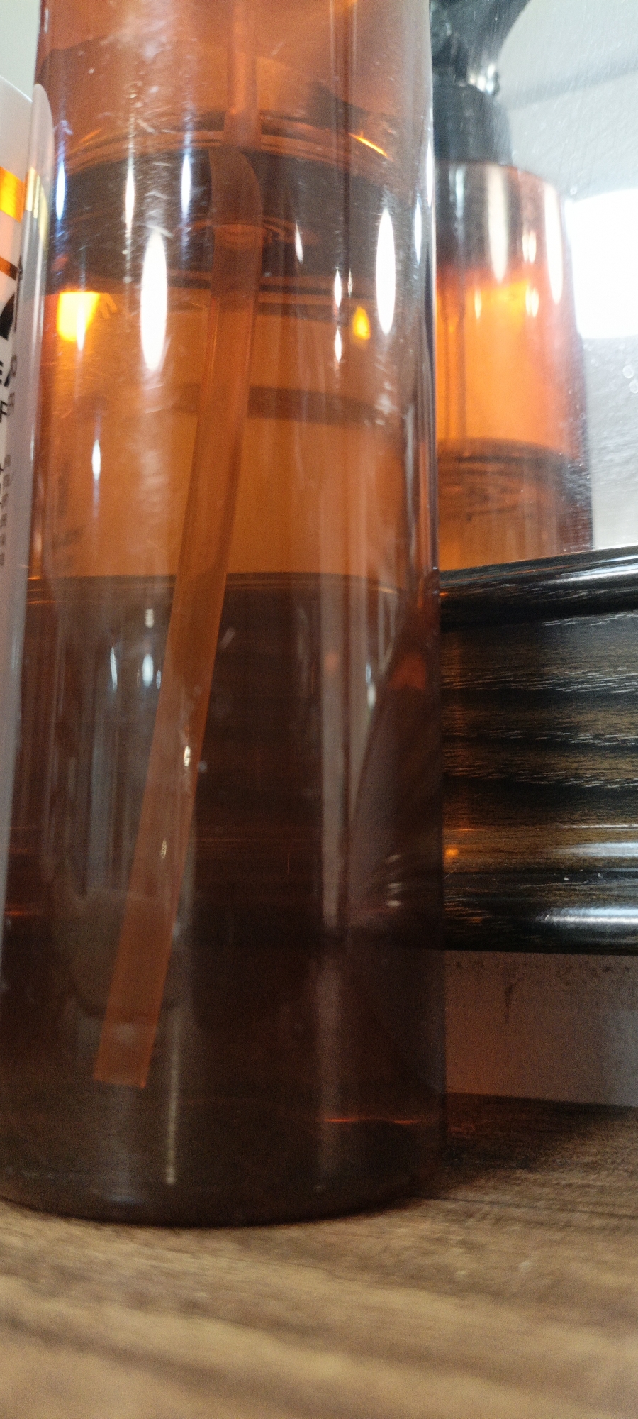 A close-up, low-angle shot shows the side of a large, clear plastic bottle with a dispensing pump. The bottle is filled with an amber-colored liquid to about half its height. Inside the bottle, a clear, thin tube extends from the pump mechanism down into the liquid. The bottle is positioned on a light-brown, wooden surface. In the background, a darker, striped object, possibly a frame or a decorative piece, is visible to the right. The lighting is warm and casts reflections on the bottle's surface, creating bright highlights. The overall impression is of a personal care or cleaning product. The scene is indoors and the specific location within a city is not determinable.